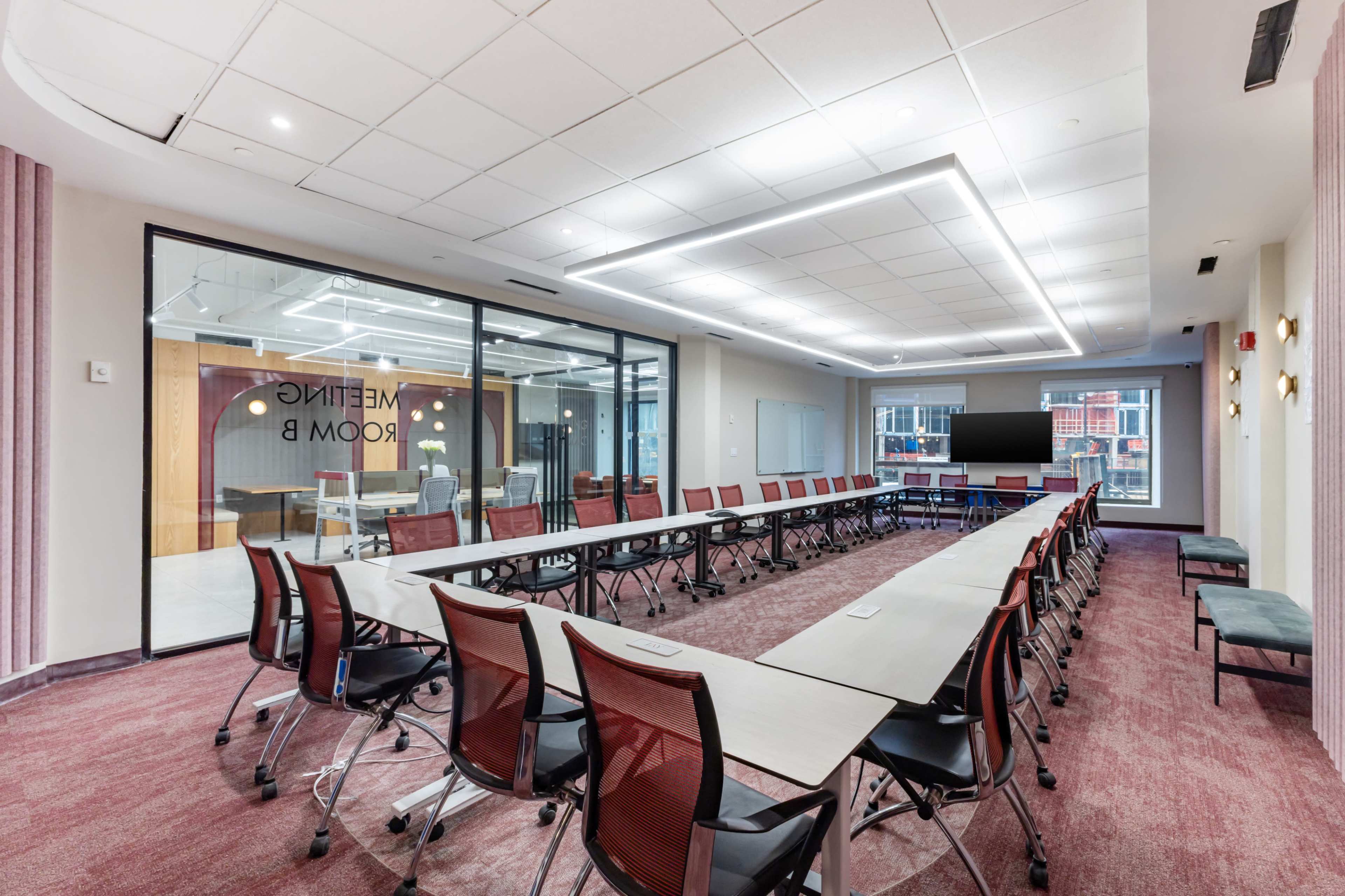 The image shows a modern meeting room arranged with numerous tables and chairs, equipped with a screen and large glass windows.