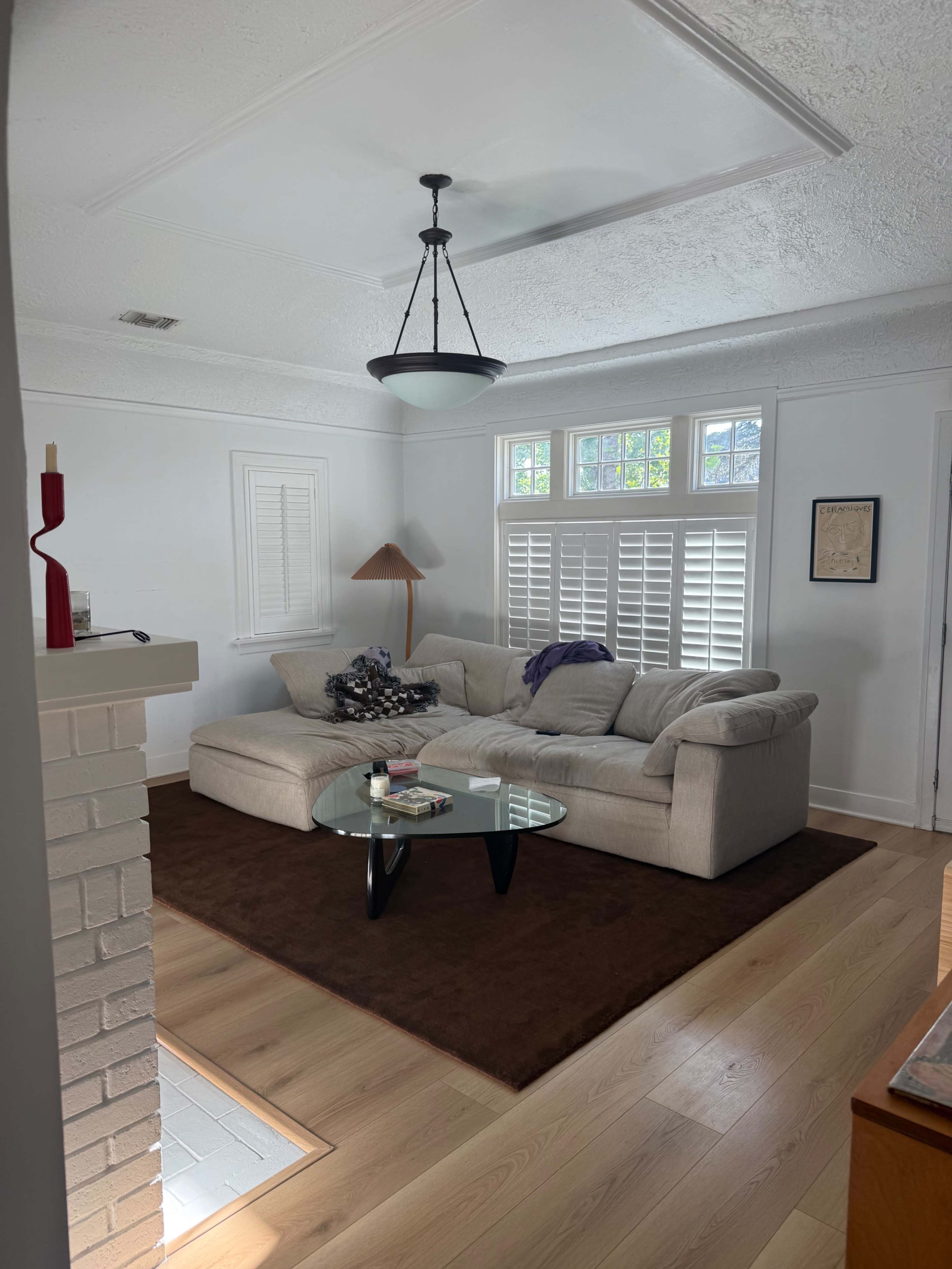 The image shows a cozy living room featuring a light-colored sectional sofa, a glass coffee table, and large windows with shutters.