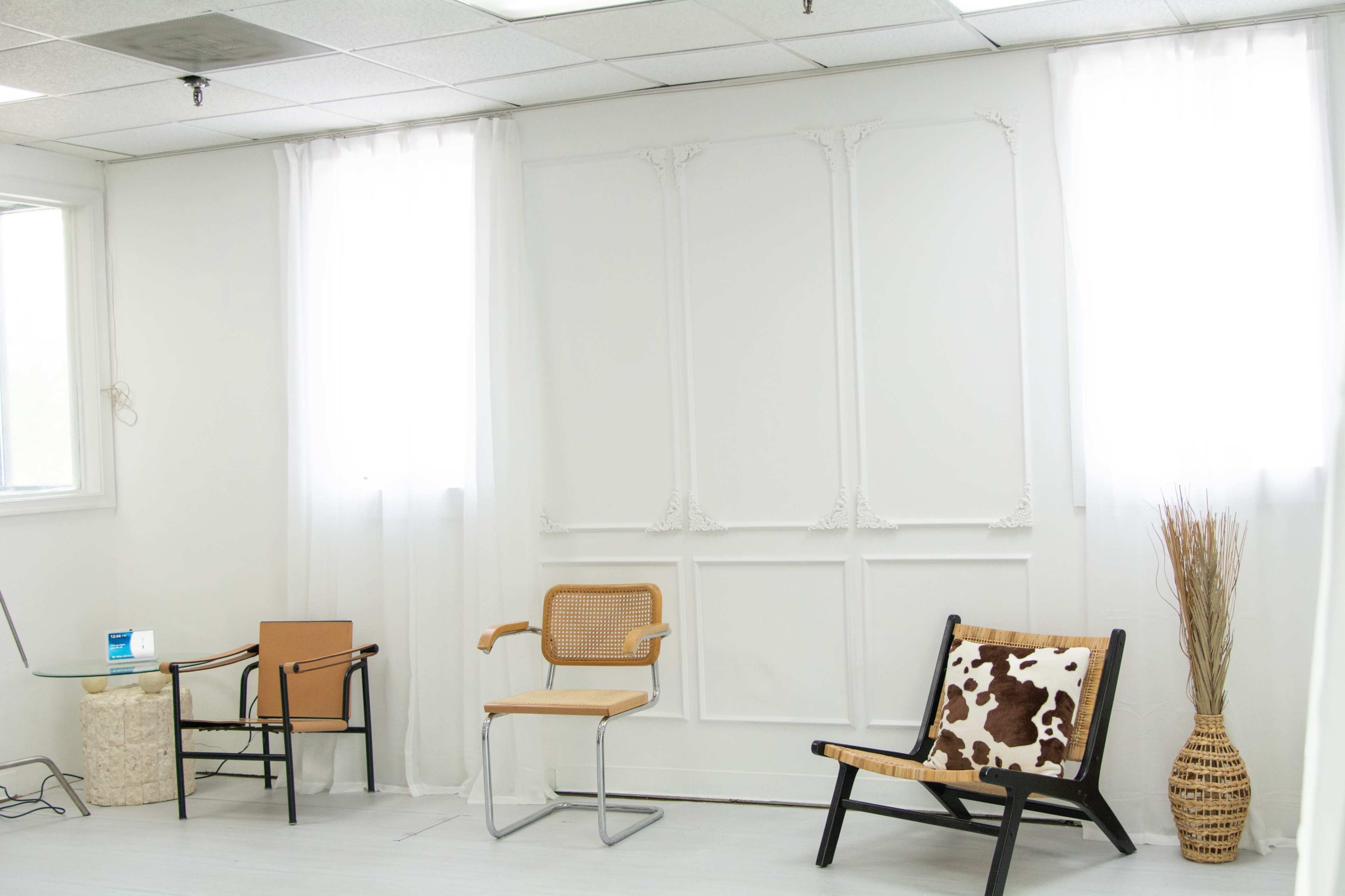 The image shows a minimalist interior space with a white wall, two large windows with sheer curtains, and a few chairs arranged around a decorative plant.
