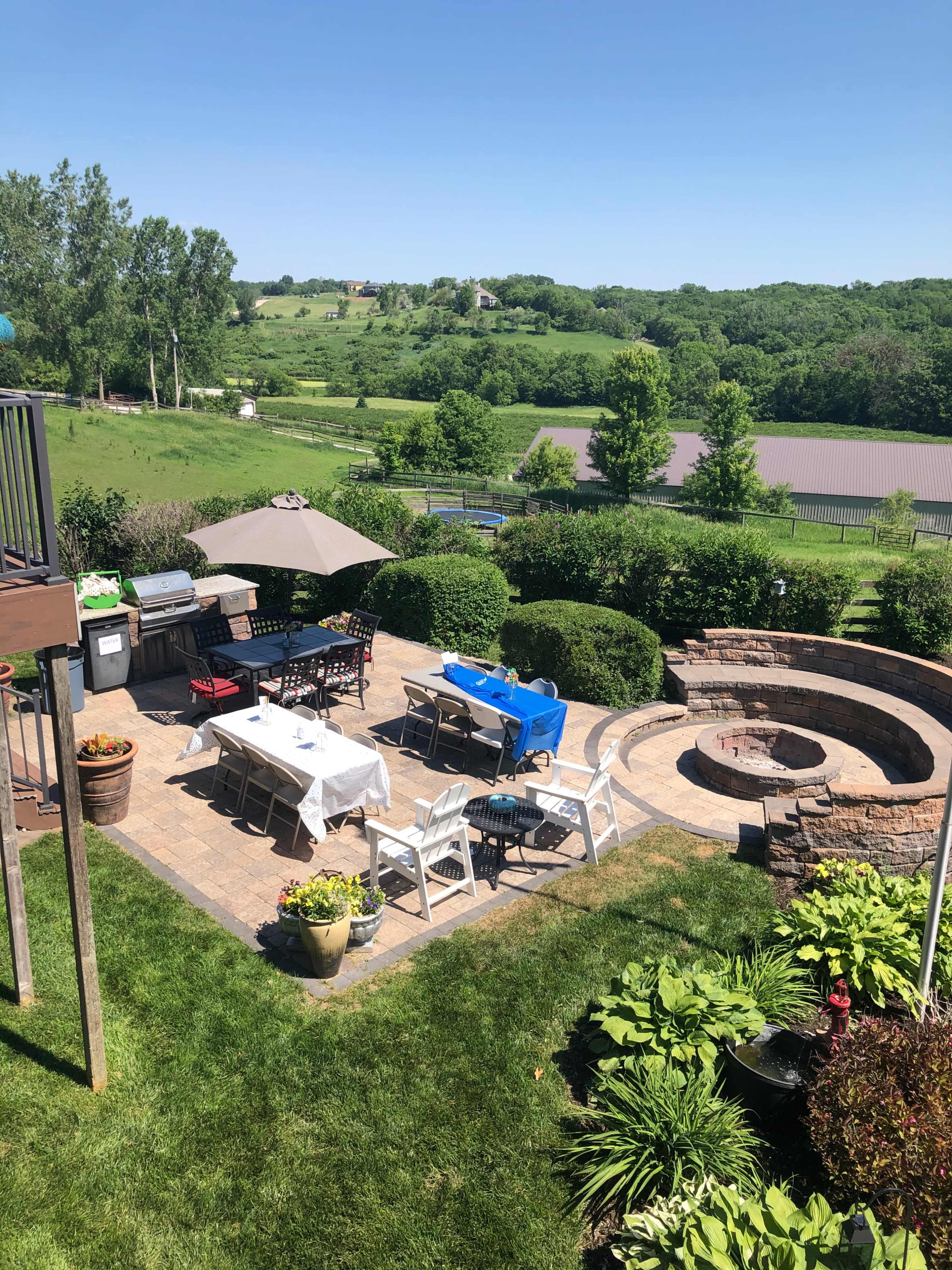 A patio with outdoor furniture and a grill is set up in a green, landscaped backyard overlooking fields and trees.
