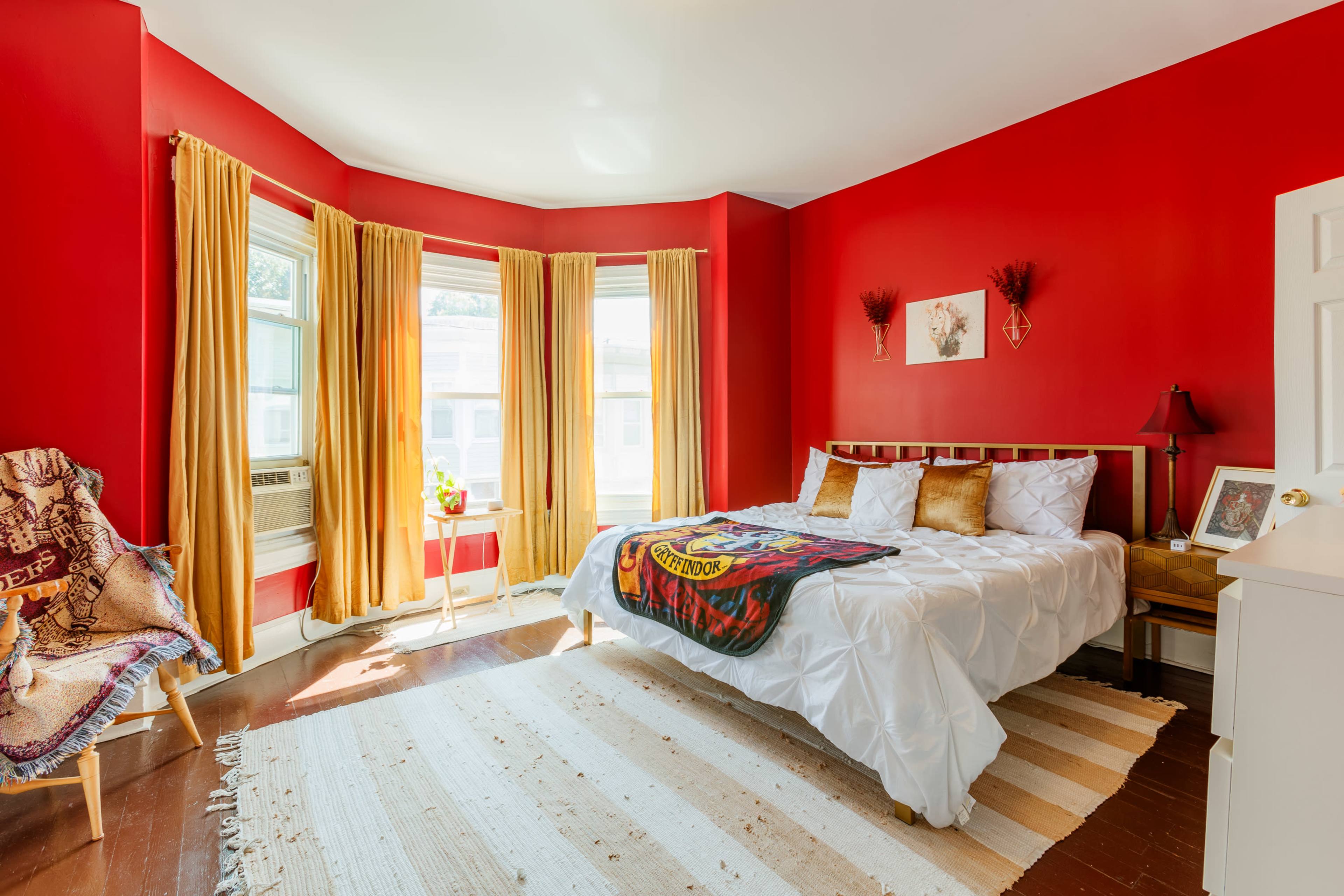The image shows a bright red bedroom with a bed, decorative pillows, a wooden nightstand, and large windows draped with yellow curtains.