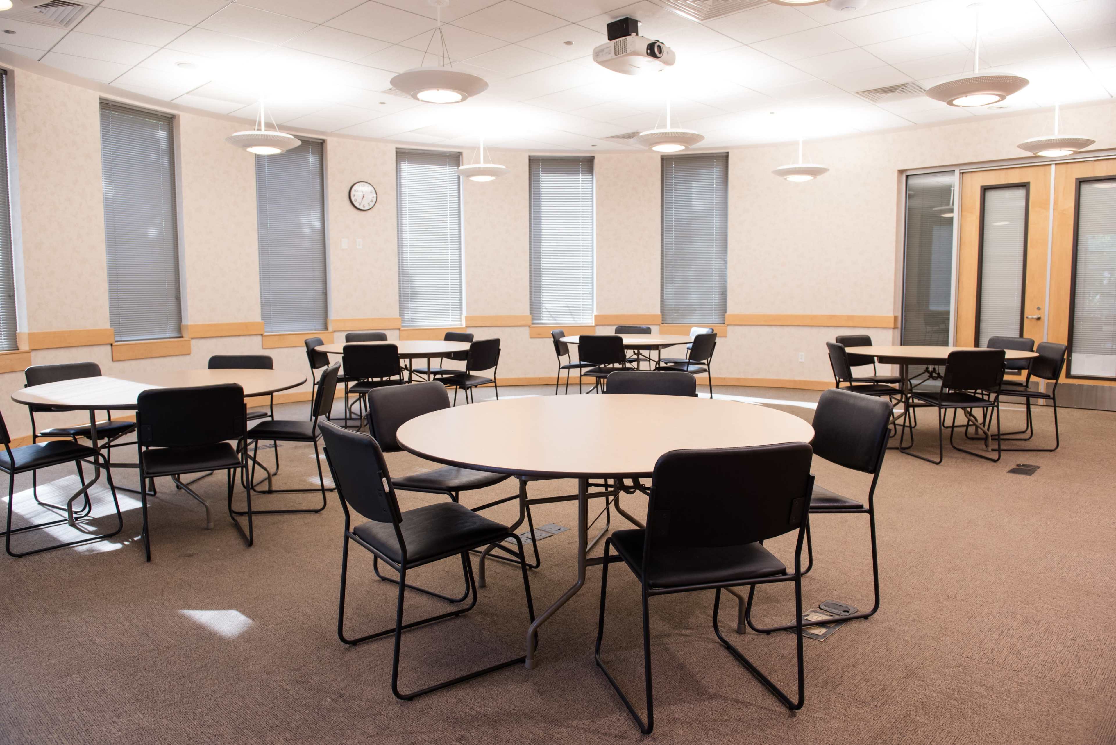 A mostly empty classroom with several round tables and black chairs arranged in a circular pattern under overhead lights.
