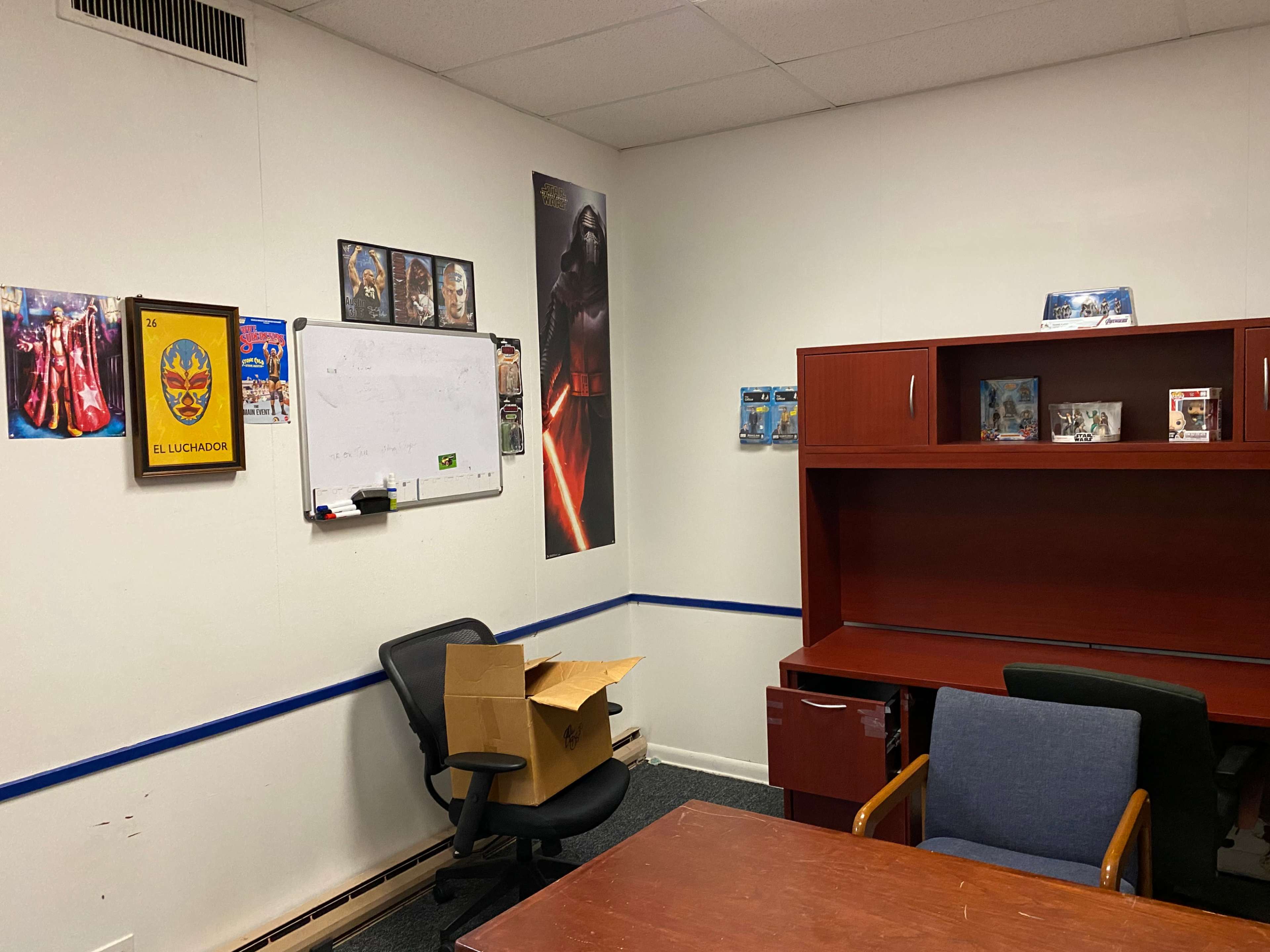 The image shows a sparsely decorated office with a whiteboard, two chairs, a wooden desk unit, a cardboard box on the floor, and several posters and items on the walls.