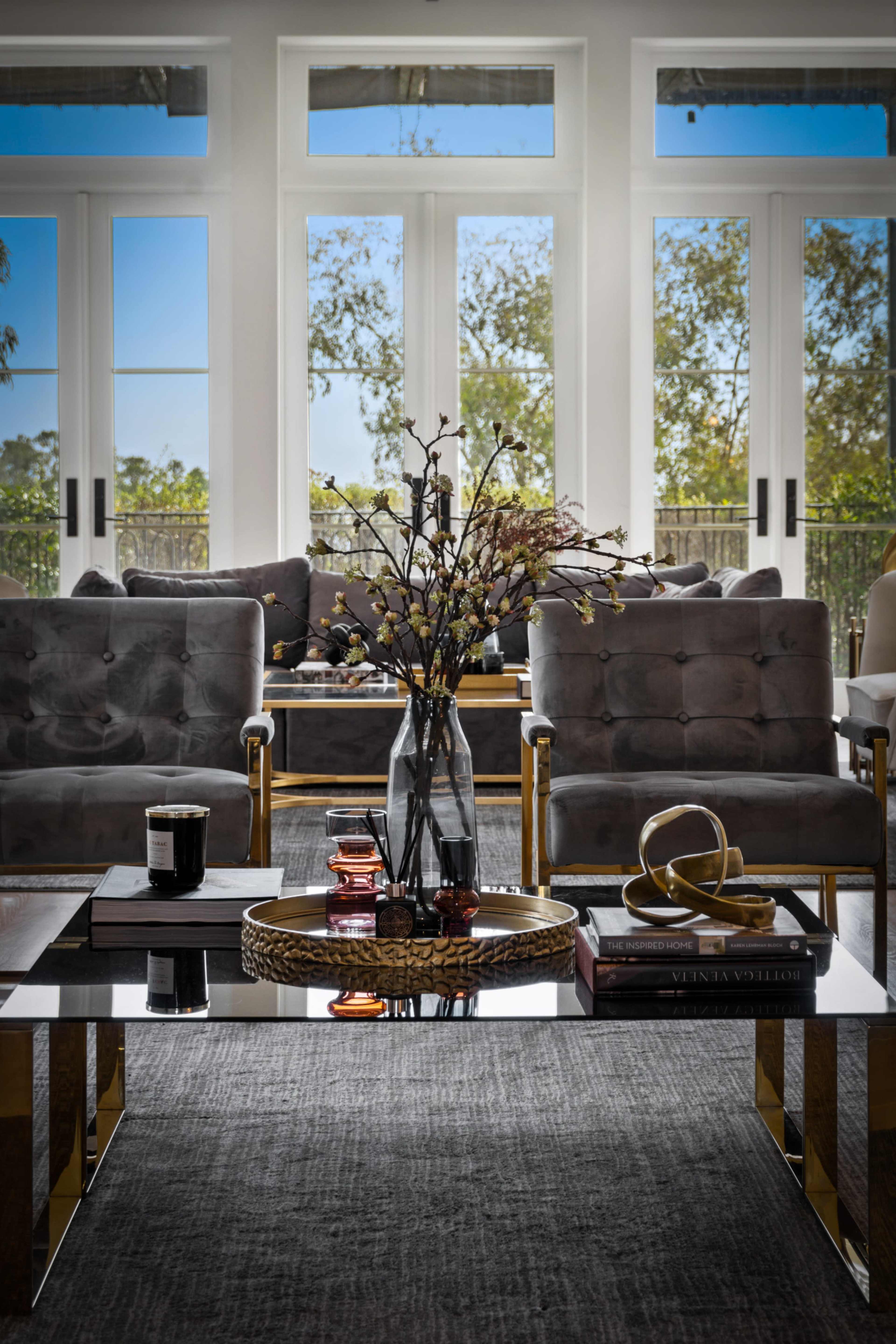 A modern living room features two gray armchairs facing a glass coffee table adorned with a vase of flowers, candles, and decorative books, with large windows providing natural light.