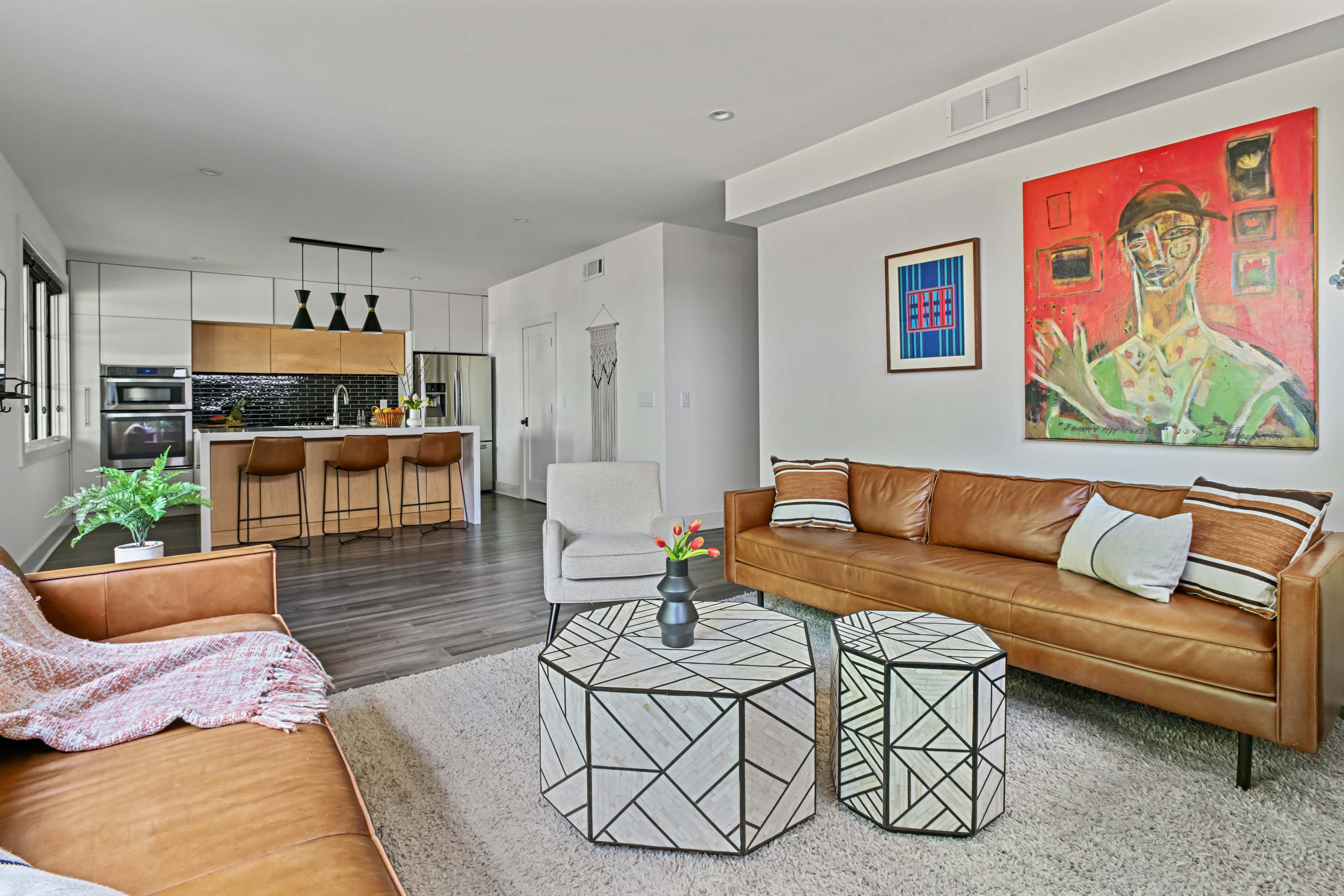 The image shows a modern living room featuring two leather sofas, geometric coffee tables, a colorful painting on the wall, and an open kitchen area in the background.