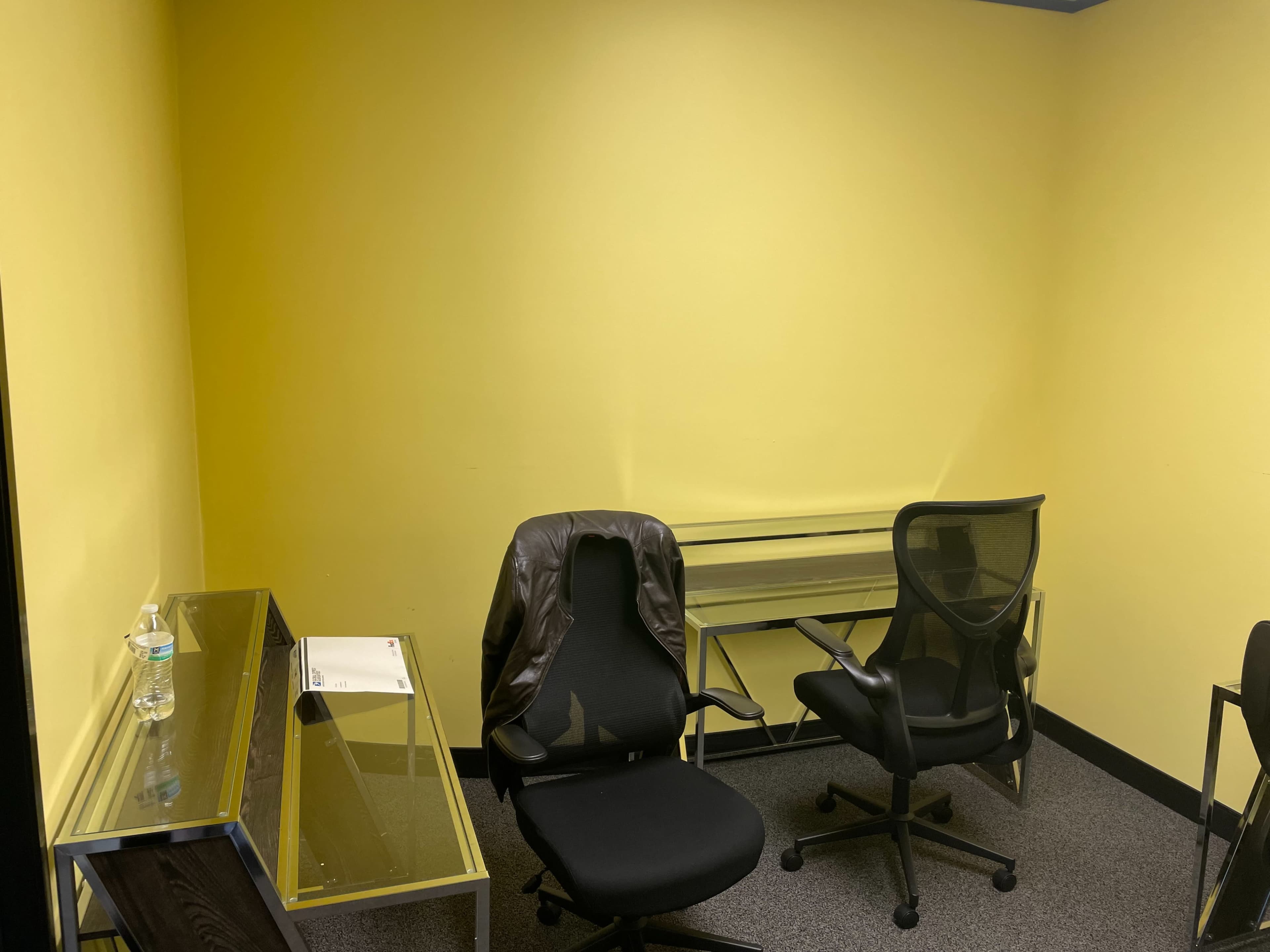 A small meeting room with yellow walls, two black office chairs, a glass desk, and a water bottle on the floor.