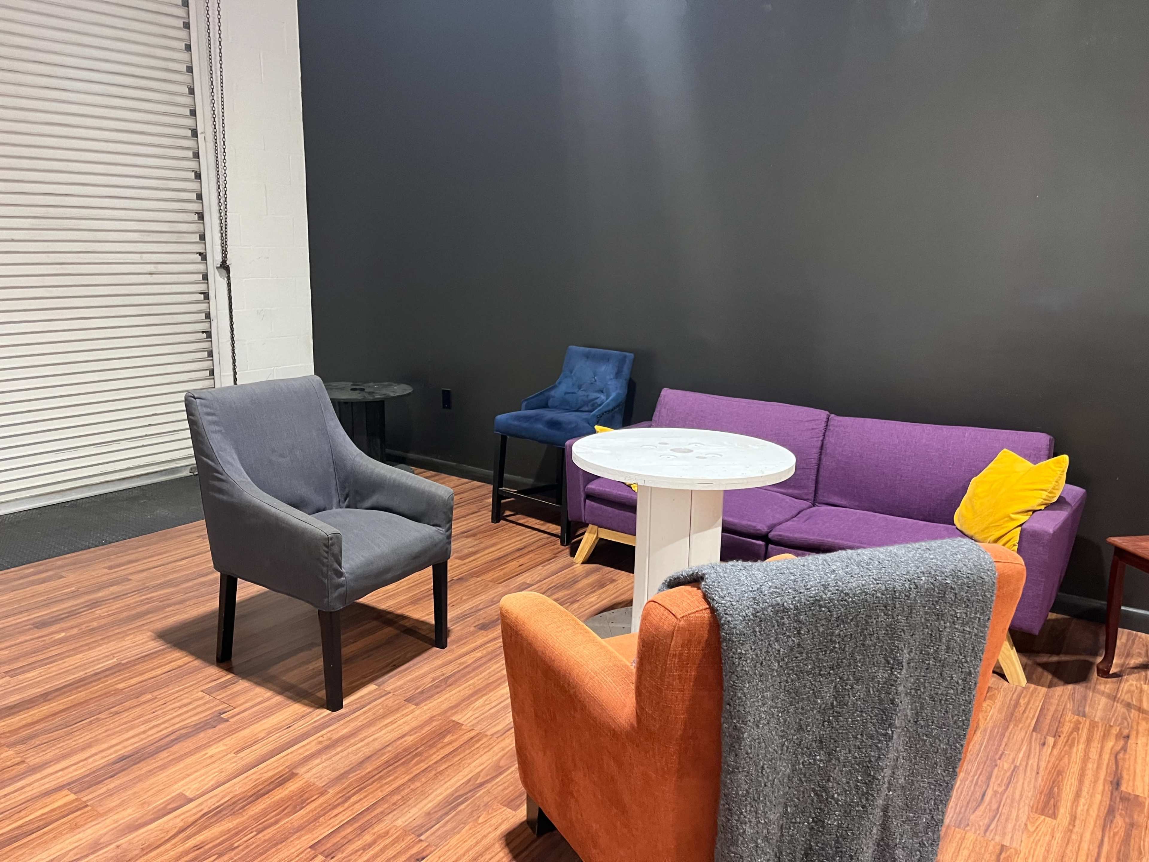 The image shows a seating area with a purple couch, an orange armchair, a gray armchair, a small white table, and a black side table against a dark wall.