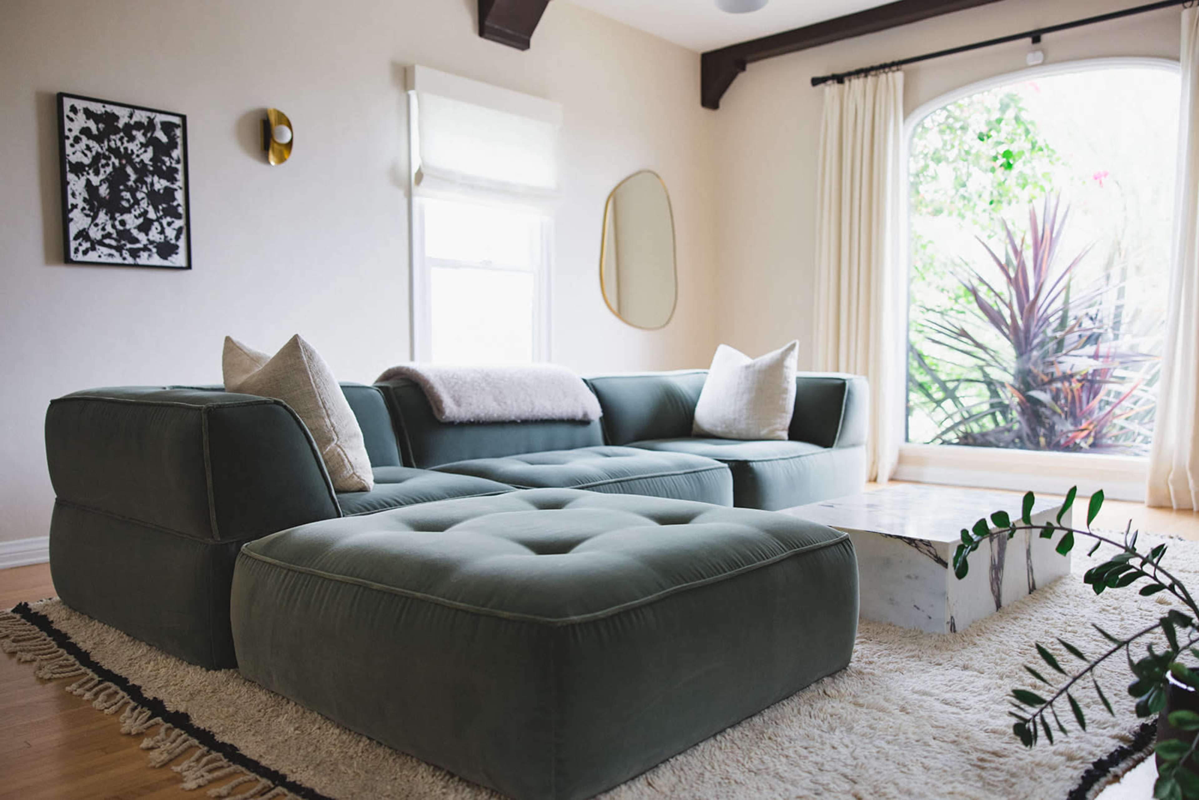 A modern living room featuring a green sectional sofa, a white marble coffee table, and large windows with natural light streaming in.