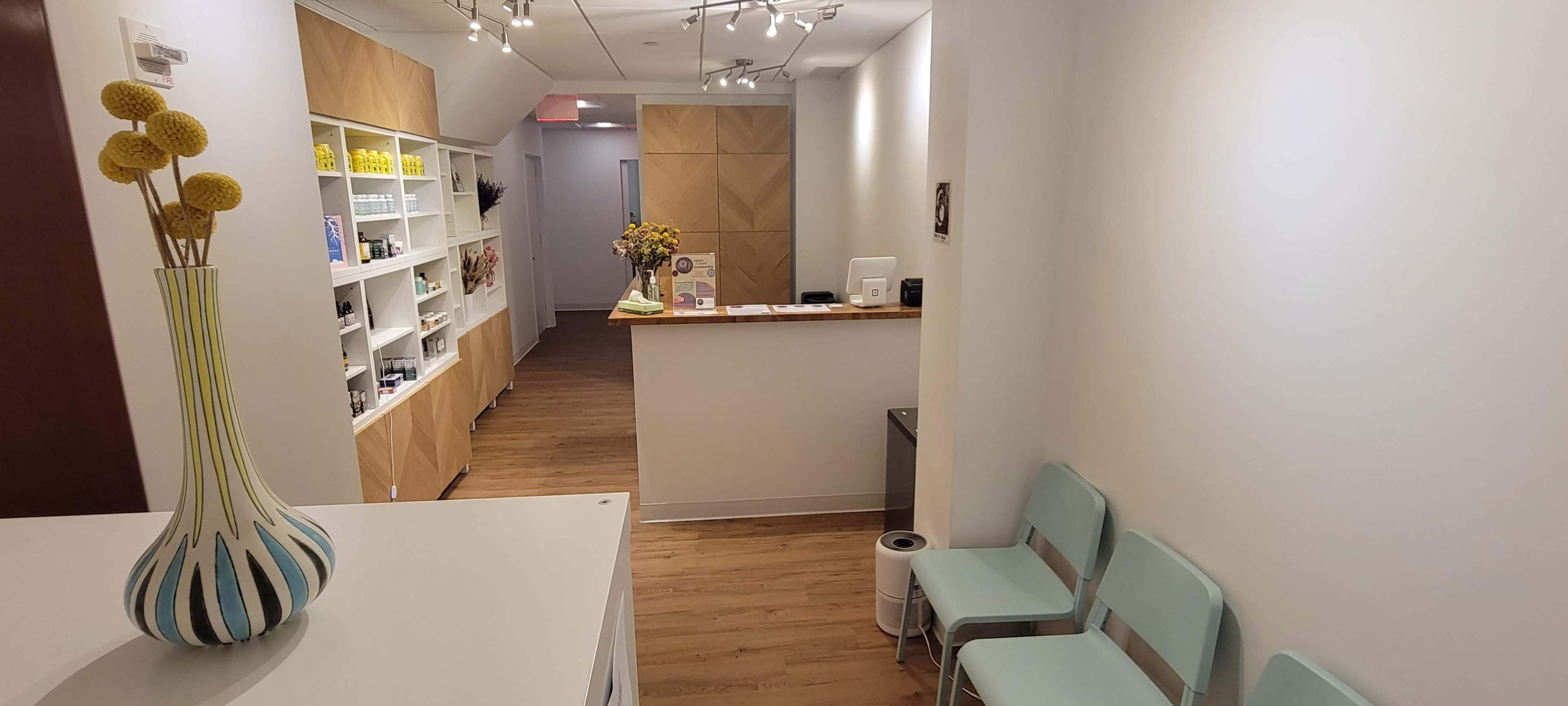 A tidy reception area with light wooden flooring, a white wall, a flower arrangement on the desk, and several teal chairs arranged in a row.
