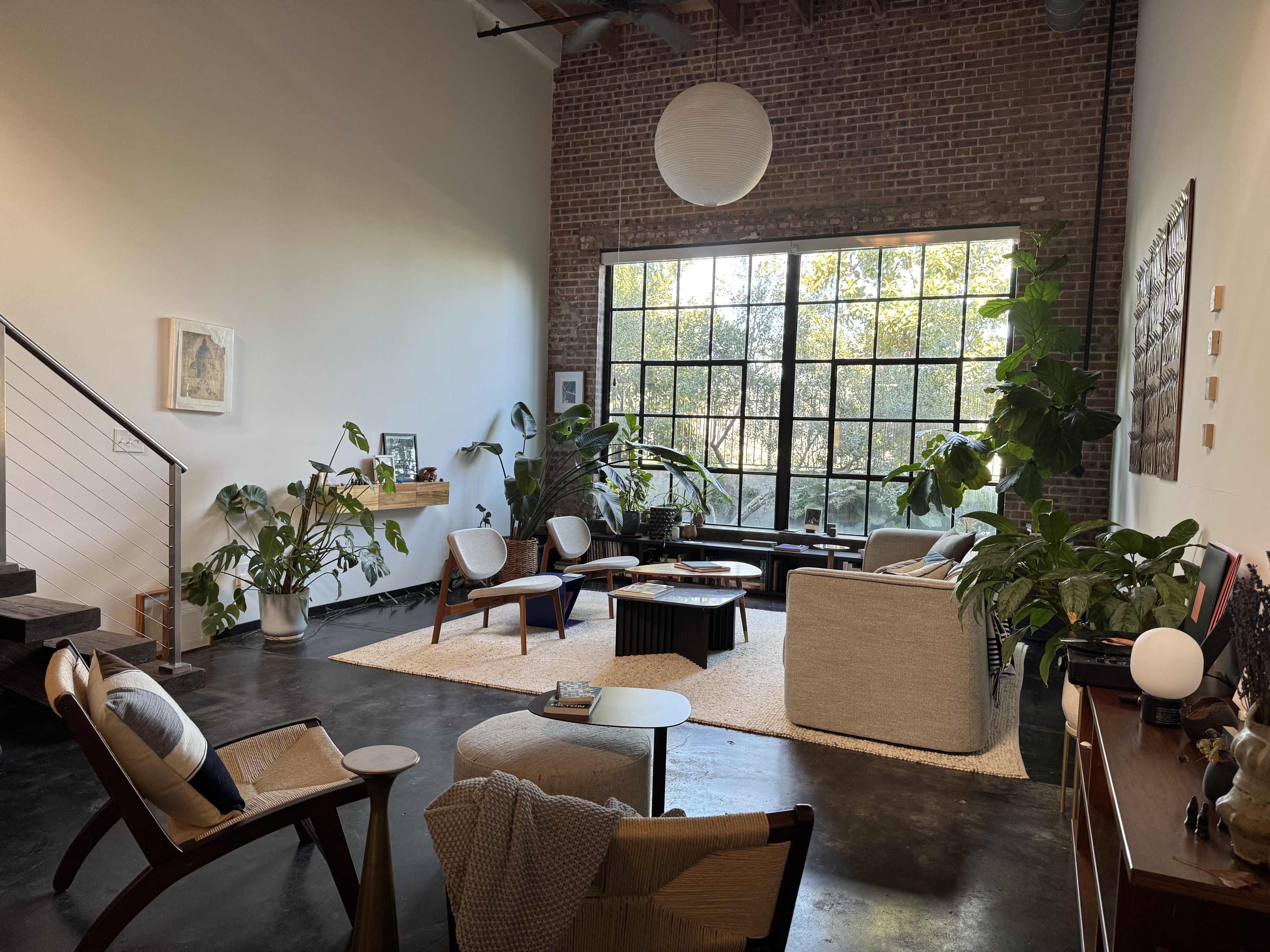 The image shows a spacious living room with large windows, modern furniture, and various indoor plants, creating a bright and inviting atmosphere.