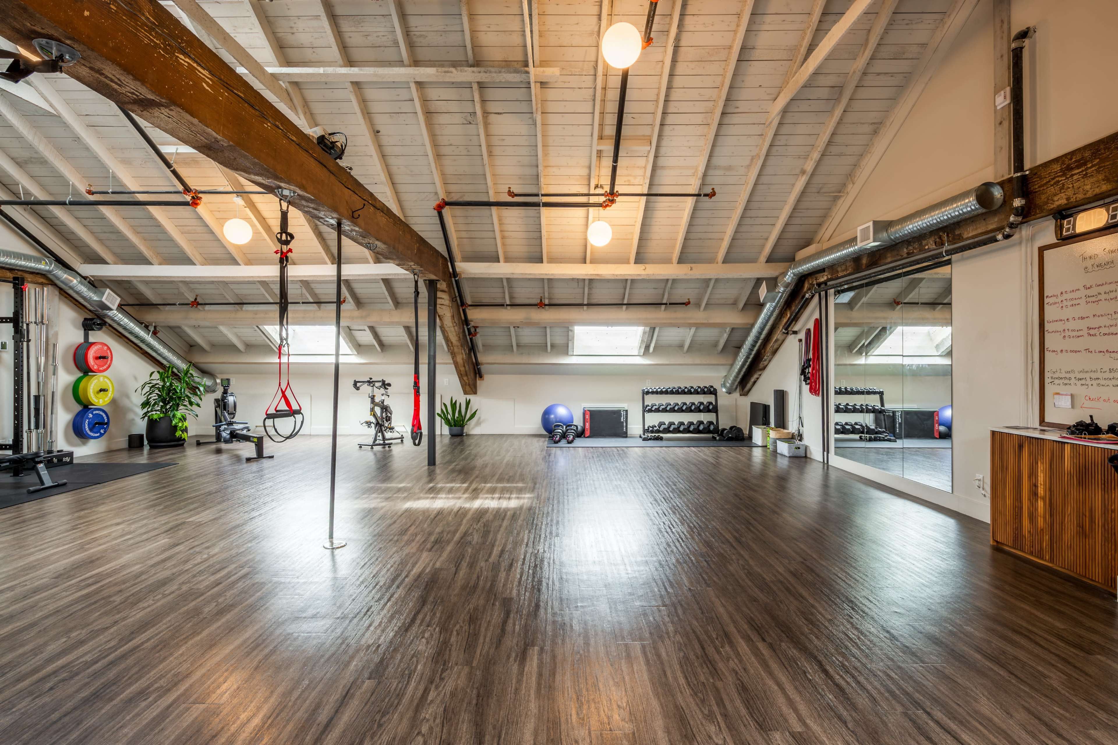 The image shows a spacious gym with wooden beams, various exercise equipment, mirrors along one wall, and colorful weights arranged in a corner.
