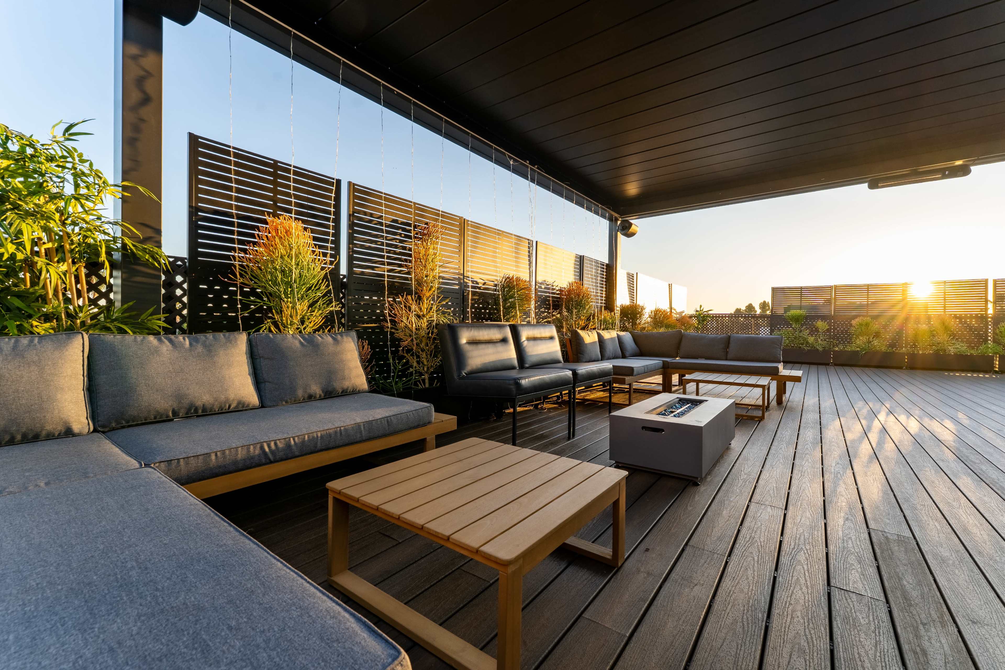 A modern rooftop terrace features comfortable seating, wooden tables, and plants, bathed in the warm light of a setting sun.