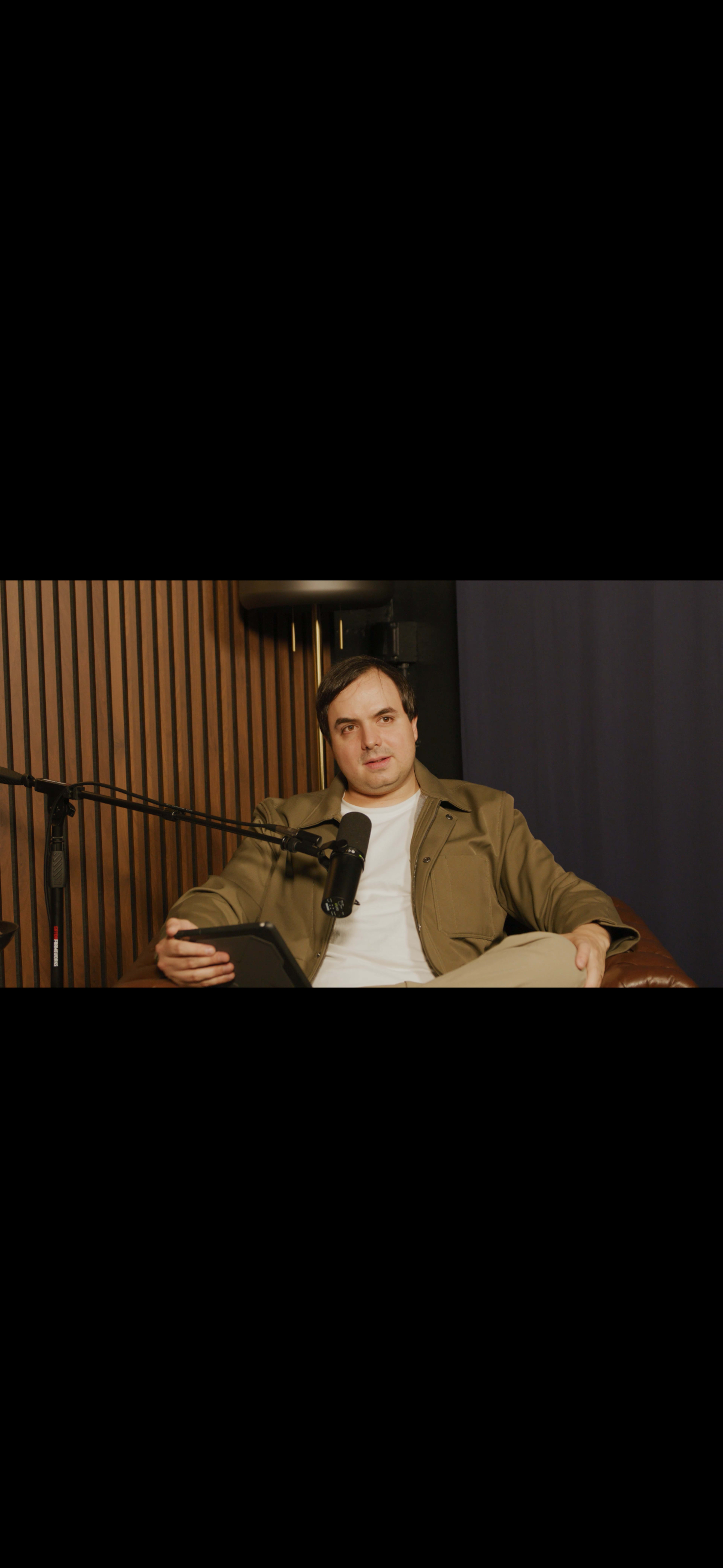 A person sits calmly in a chair, holding a device, with microphones positioned nearby in a studio setting.