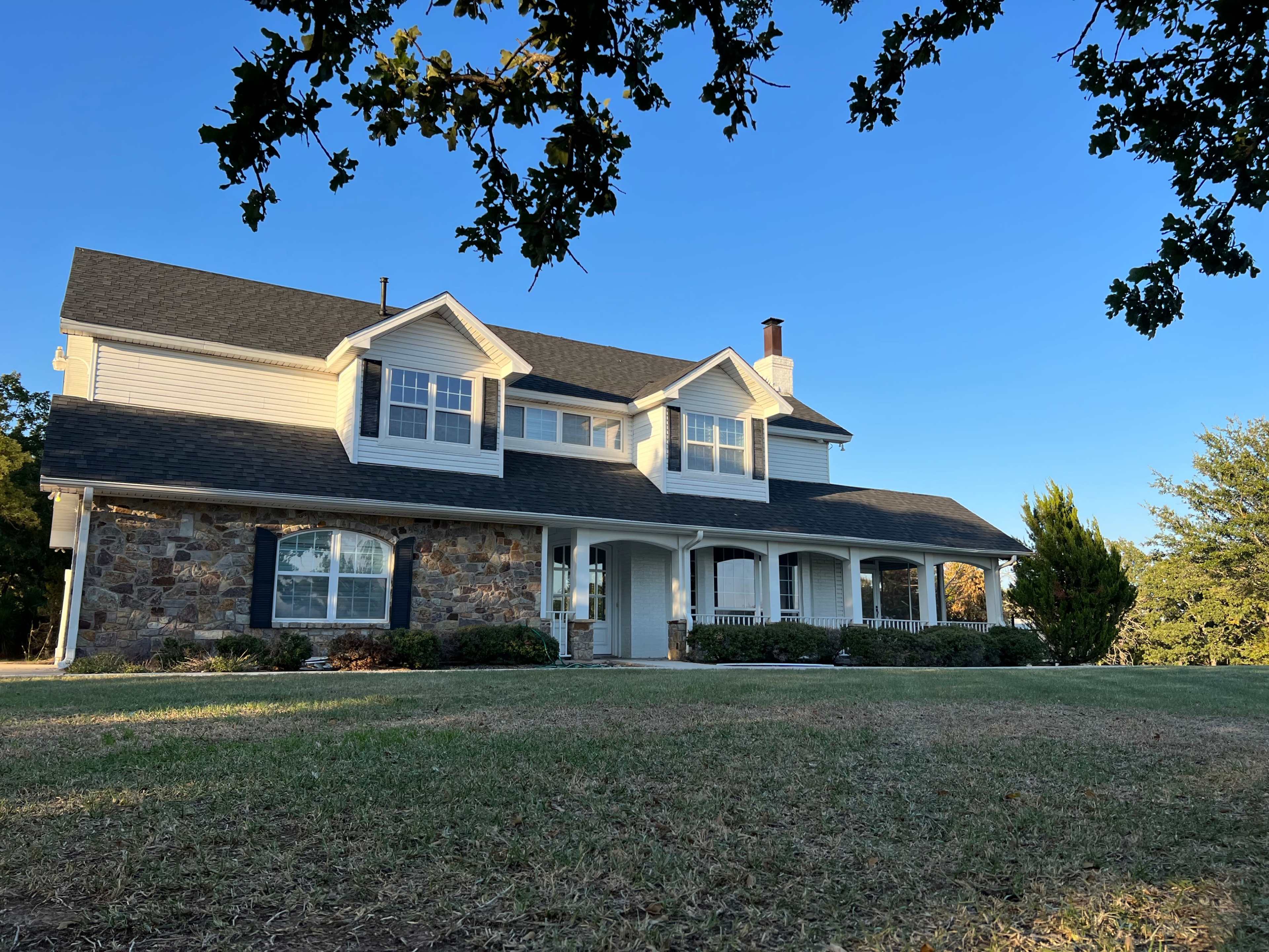 The image shows a two-story house with a stone facade and a large front porch, surrounded by a grassy lawn and trees under a clear blue sky.