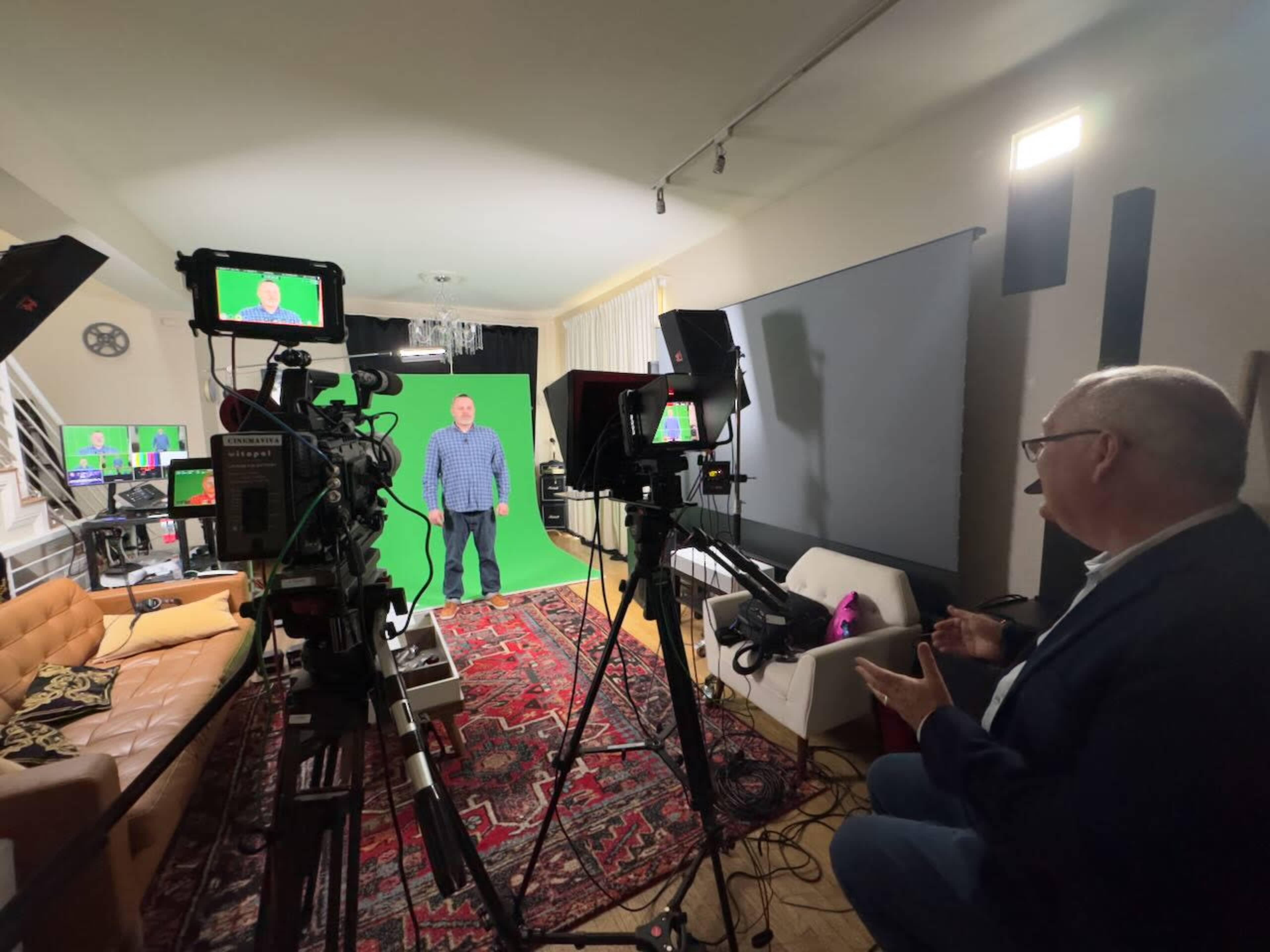 The image shows a studio setup with two cameras focused on a person standing in front of a green screen, while another individual sits nearby, possibly directing or interacting.