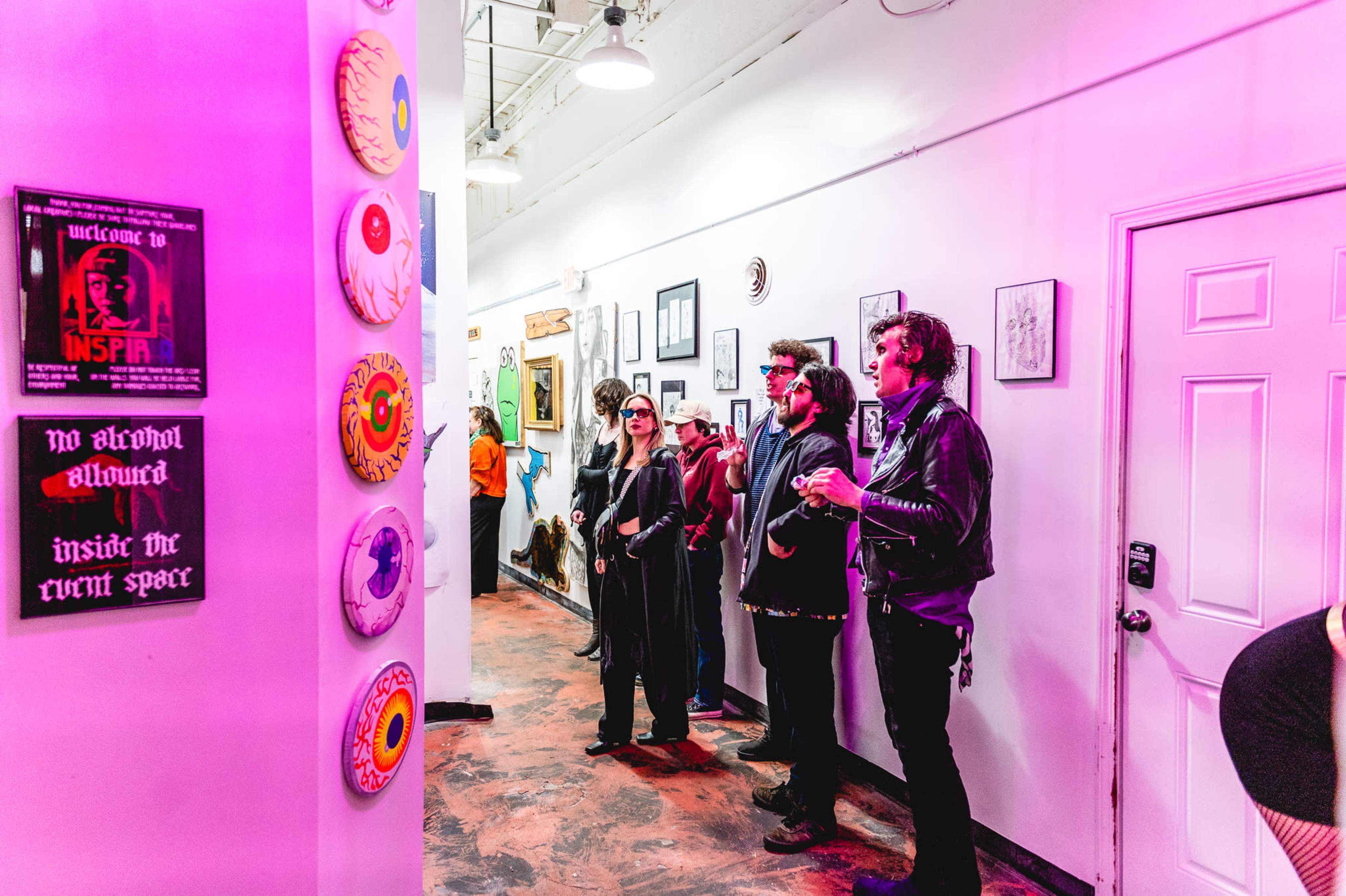 A group of three people in dark clothing stands in a brightly lit hallway adorned with colorful artwork and decorative plates.