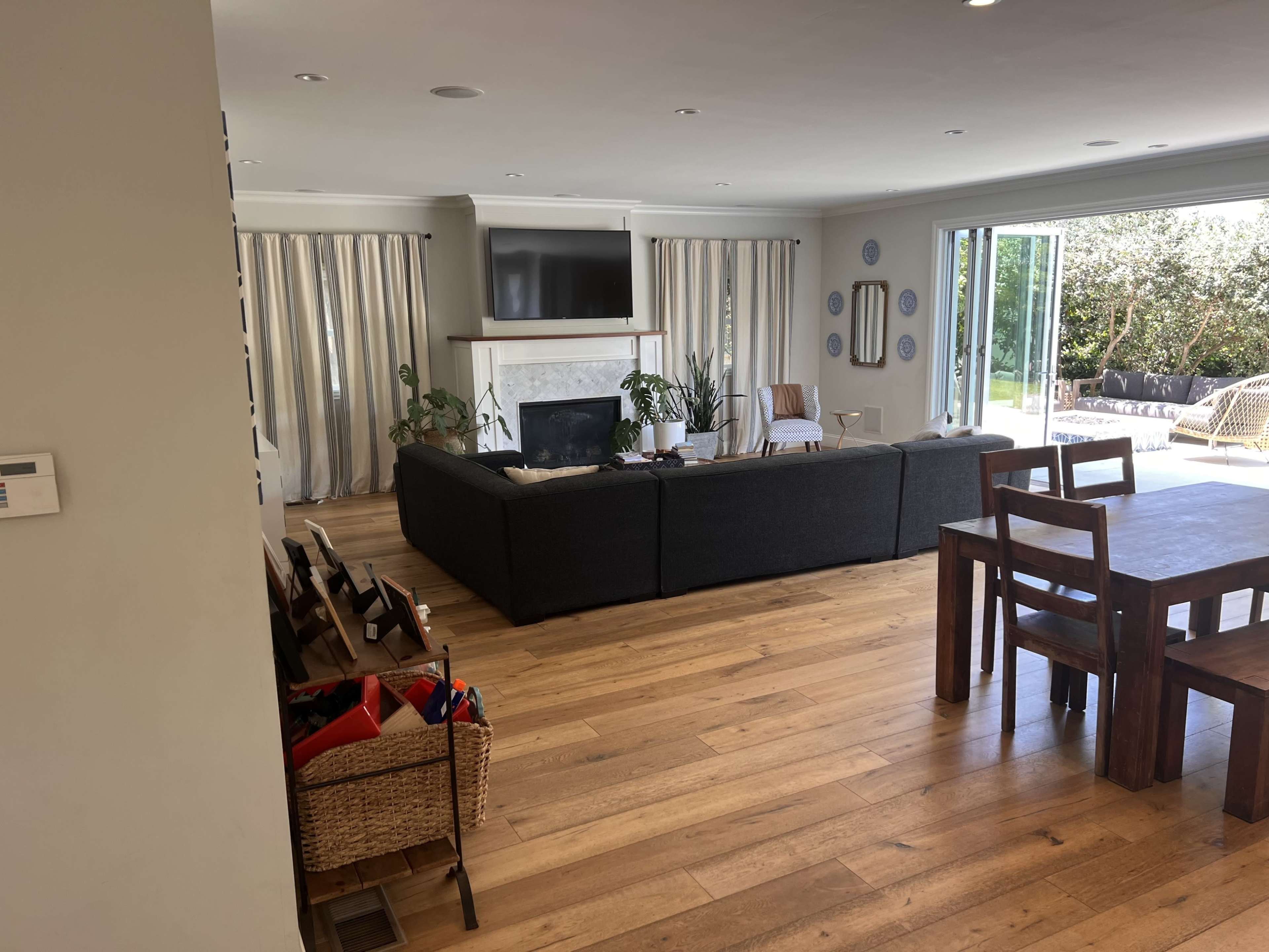A spacious living area with a dark sofa, a television above a fireplace, and large windows opening to a garden, accompanied by a dining table and wooden flooring.