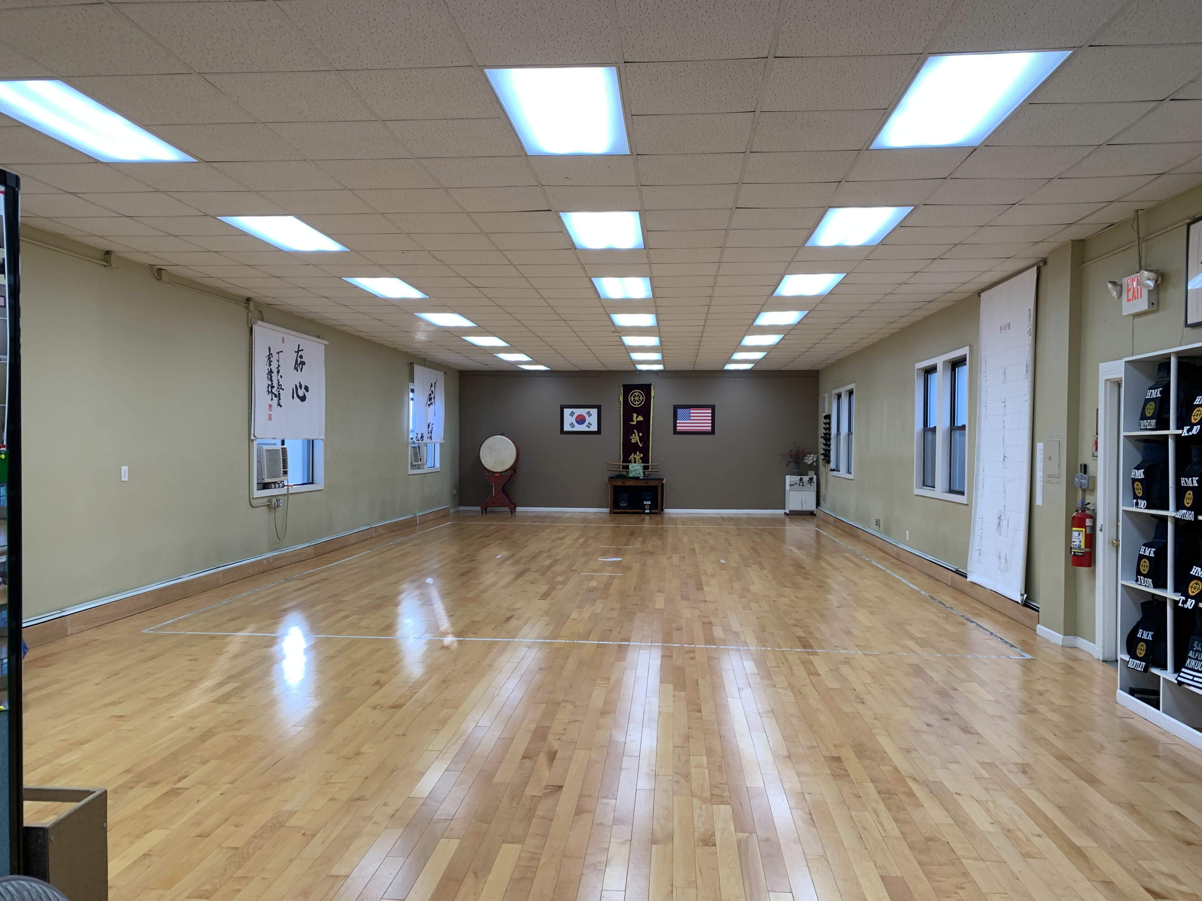 The image shows a spacious martial arts dojo with wooden flooring, wall-mounted decorations, and a small platform displaying flags and a drum.