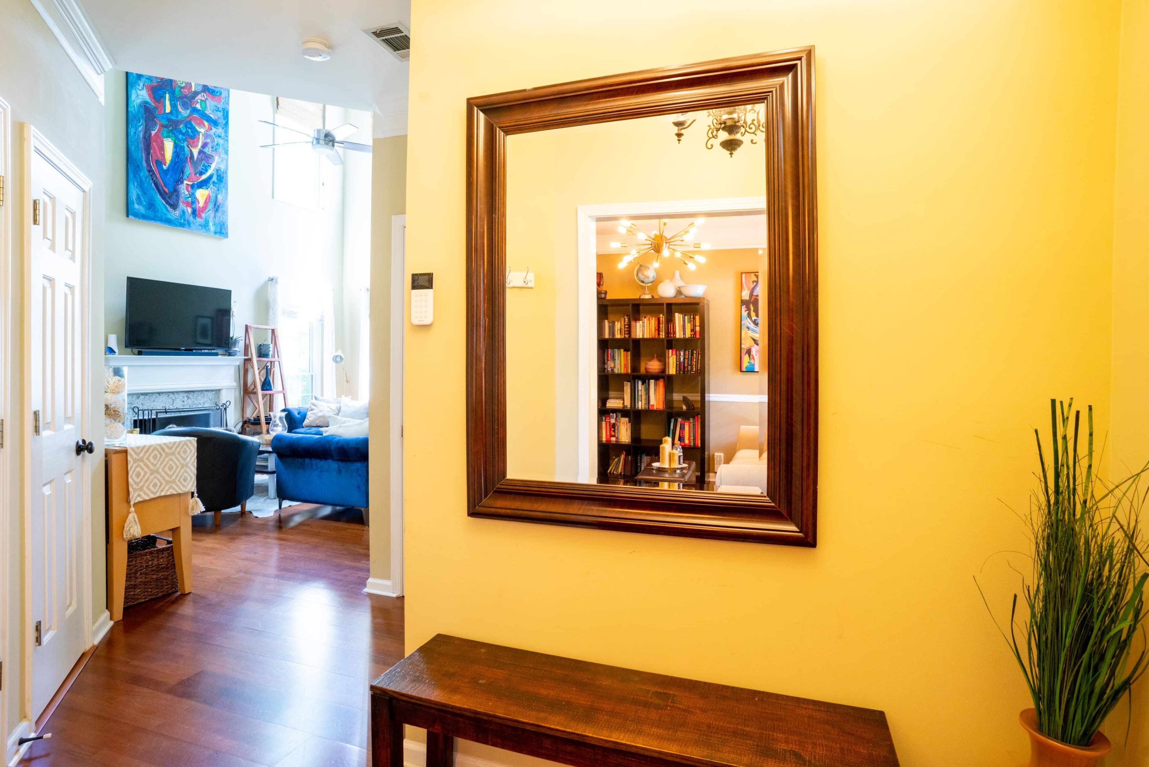 The image shows a hallway with a large mirror on the wall, a wooden bench in front, and a view into a living room featuring bookshelves and a TV in the background.