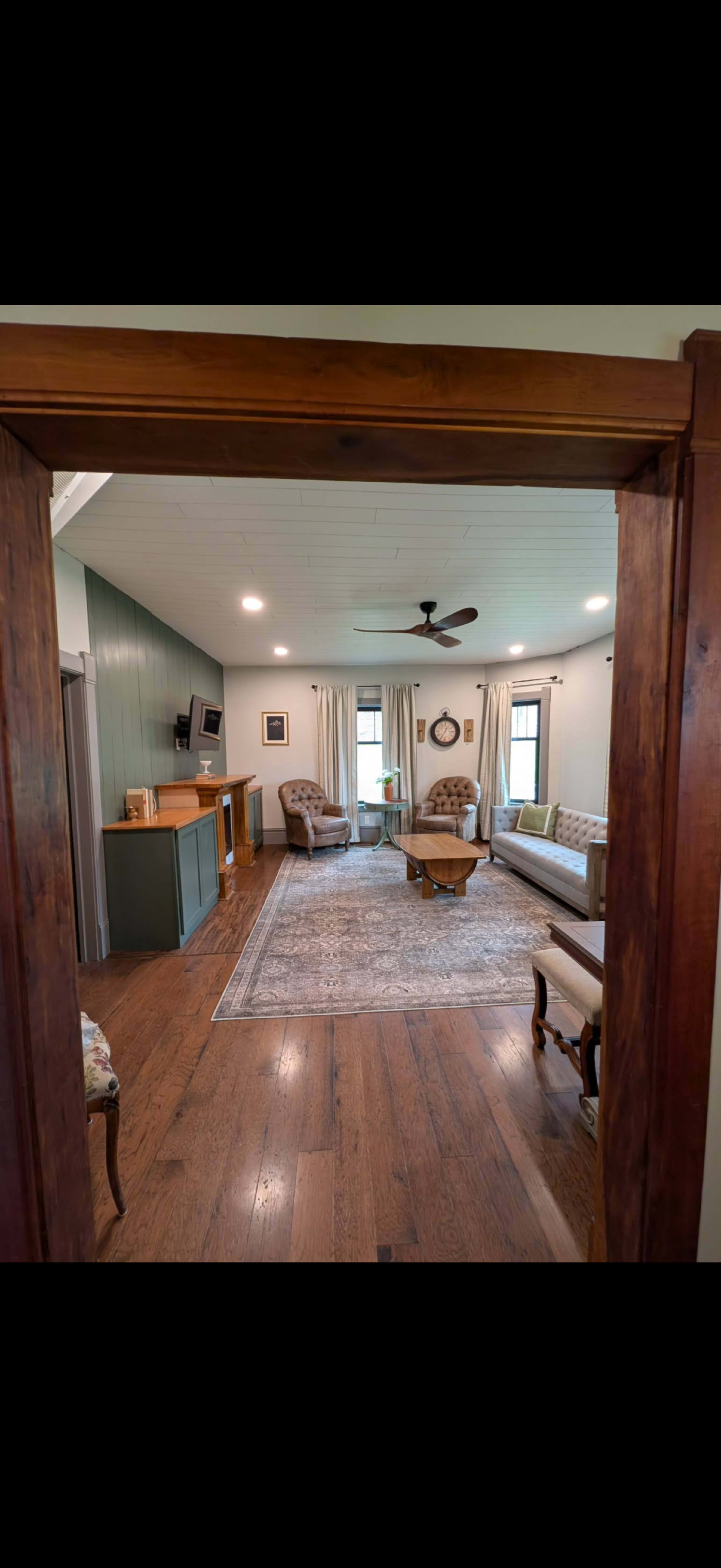 The image shows a living room with wooden flooring, a patterned area rug, and two sofas arranged around a coffee table, with a television mounted on the wall and a side console in the background.