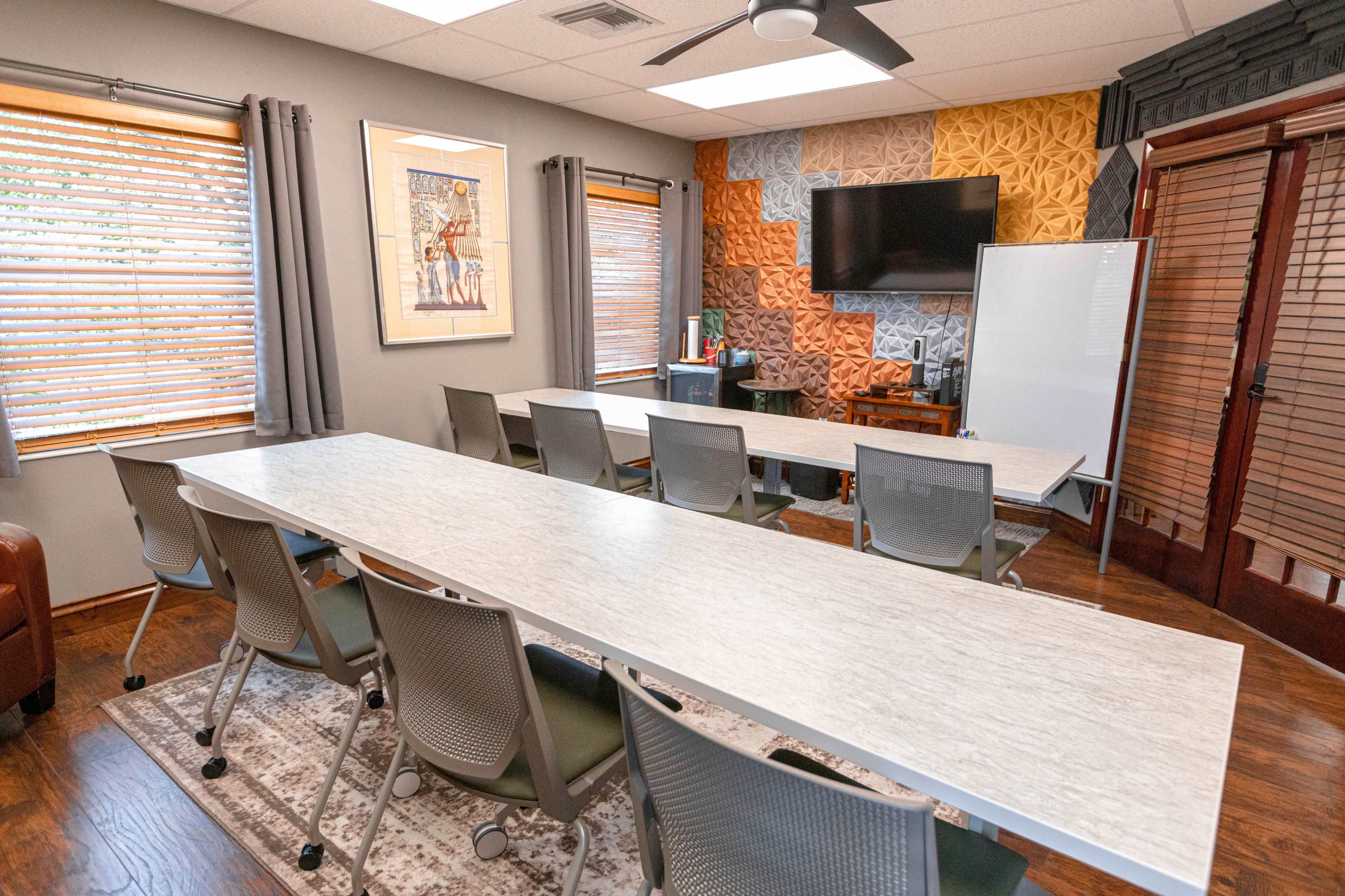 A spacious conference room features a long table with chairs, large windows with blinds, and a TV mounted on an accent wall.