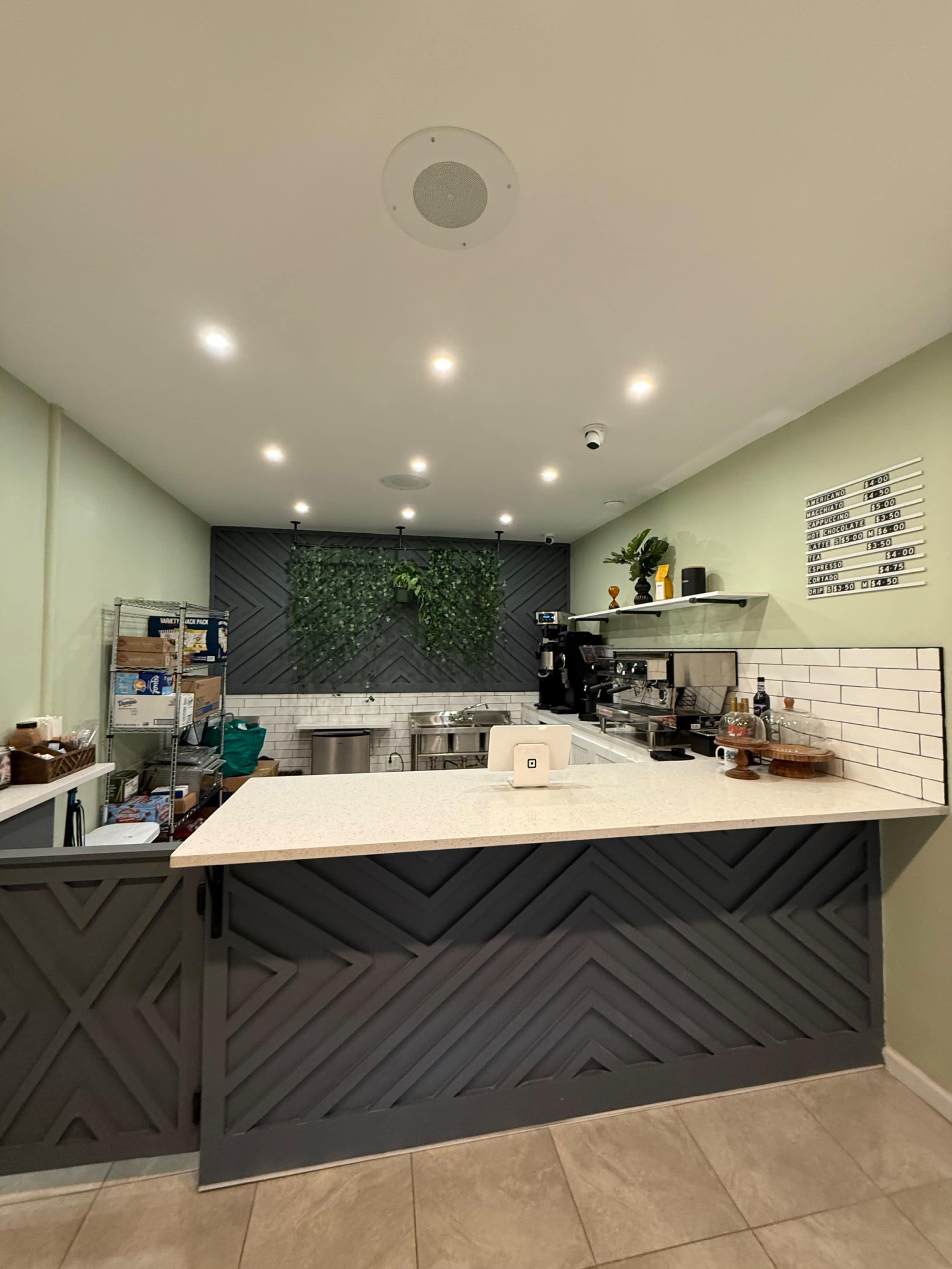 The image shows a modern kitchen space with a gray and white color scheme, featuring an island counter, shelving with kitchen equipment, and a green plant wall accent.