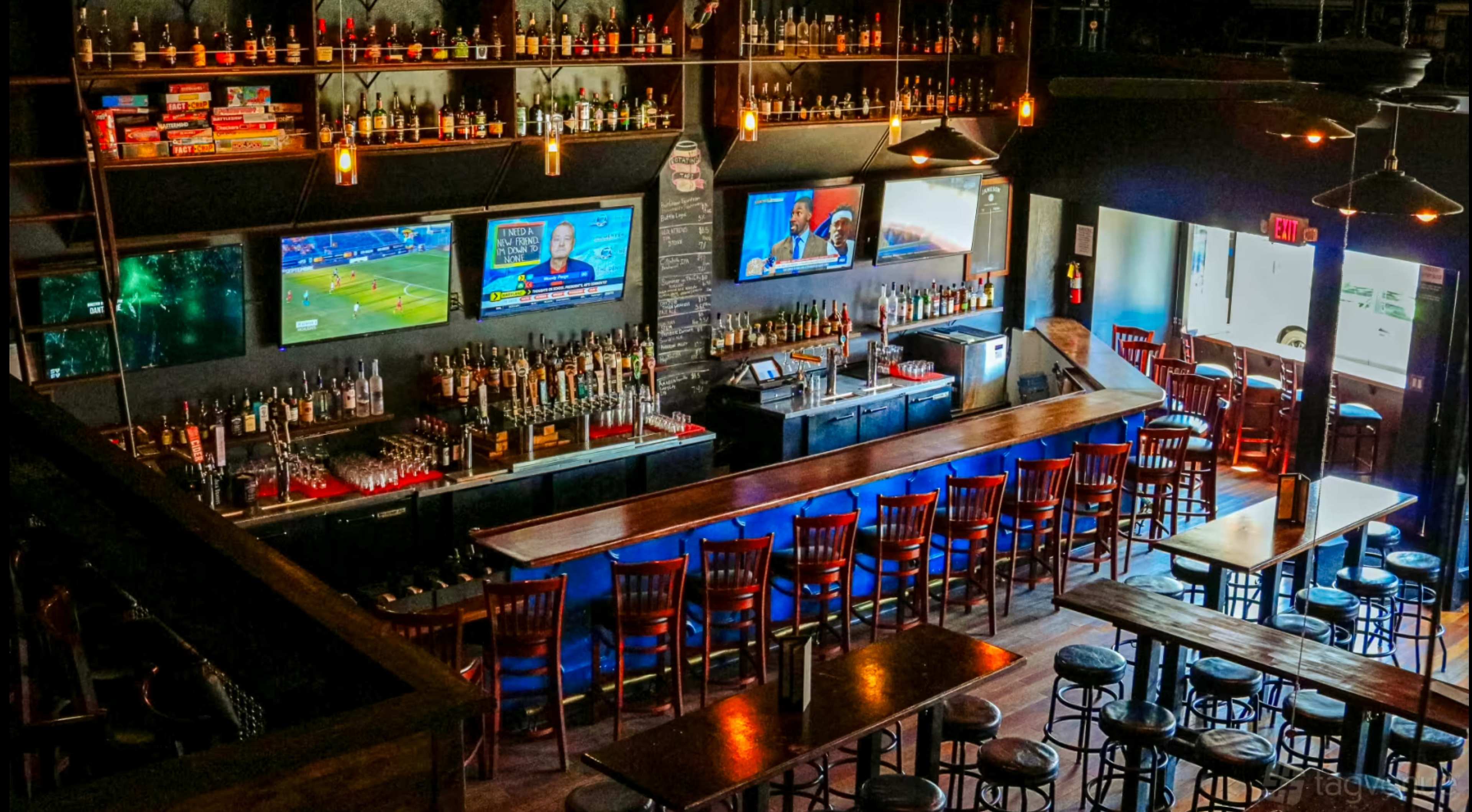 The image shows a spacious bar area with a wooden counter, multiple rows of chairs, and two large screens displaying different sports events.