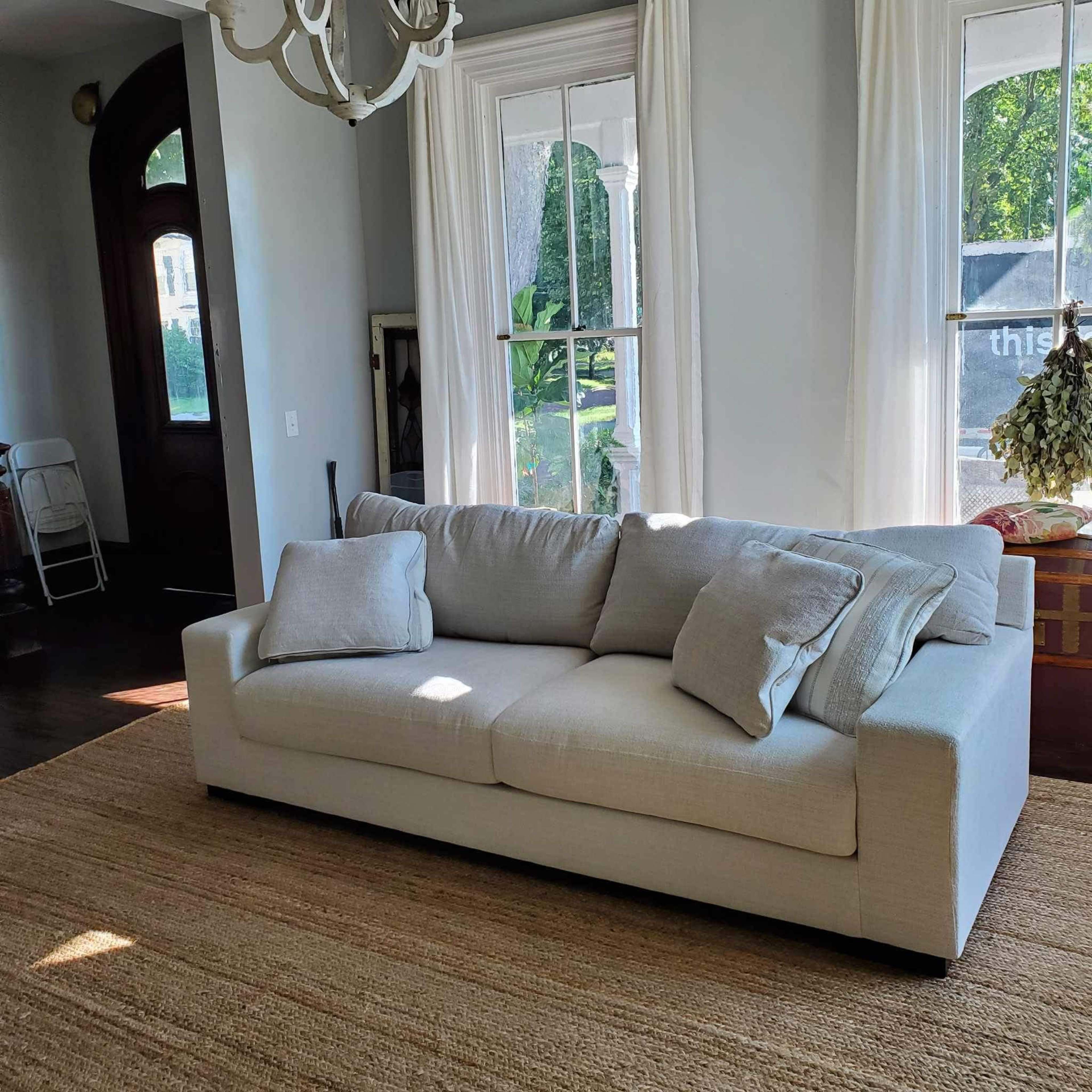 A light-colored sofa with two cushions, placed on a woven rug in a room with large windows and a doorway.