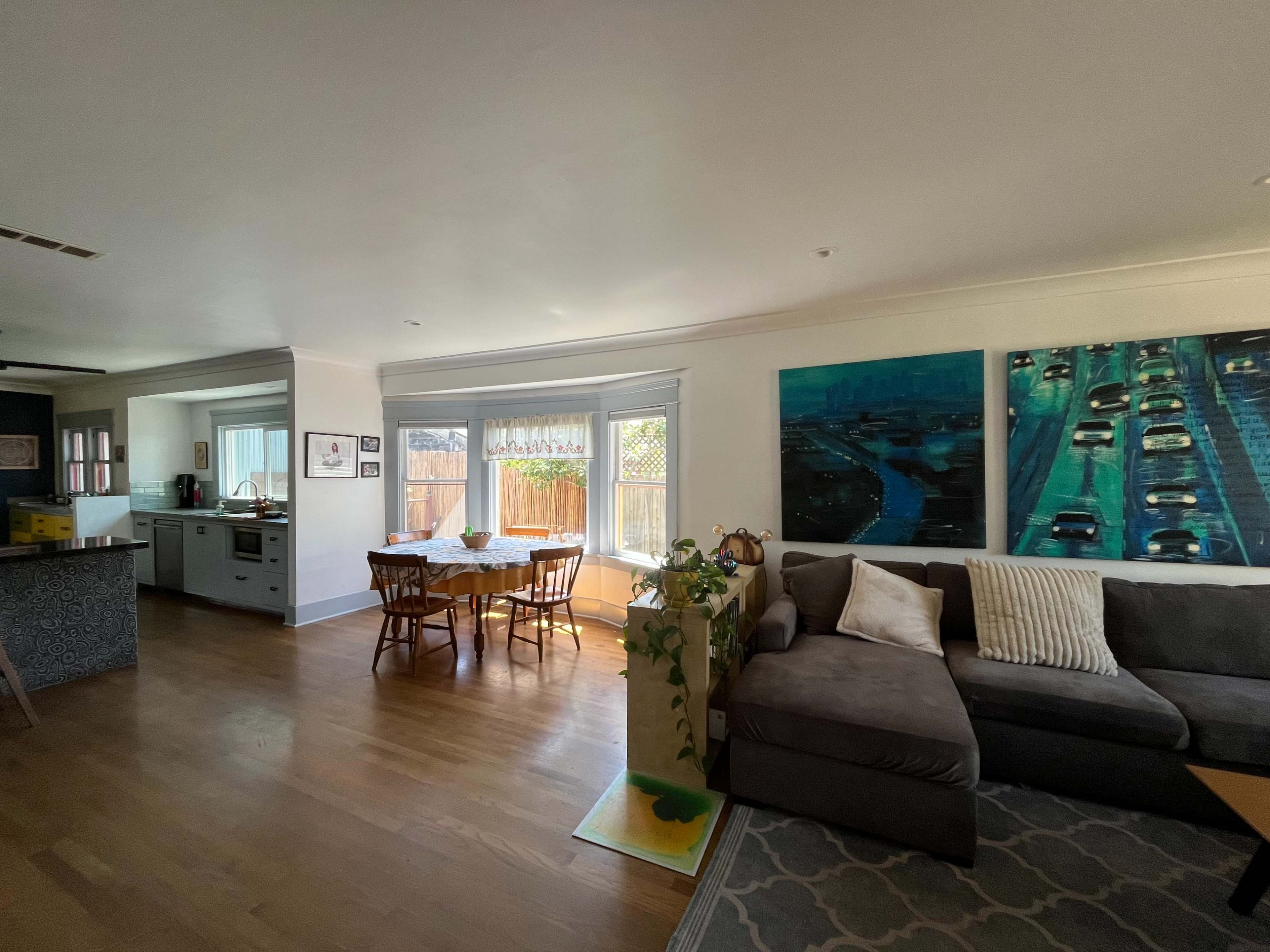 The image shows a living room with a sectional sofa, a dining area featuring a round table and chairs, and large windows allowing light into the space, with kitchen access visible in the background.