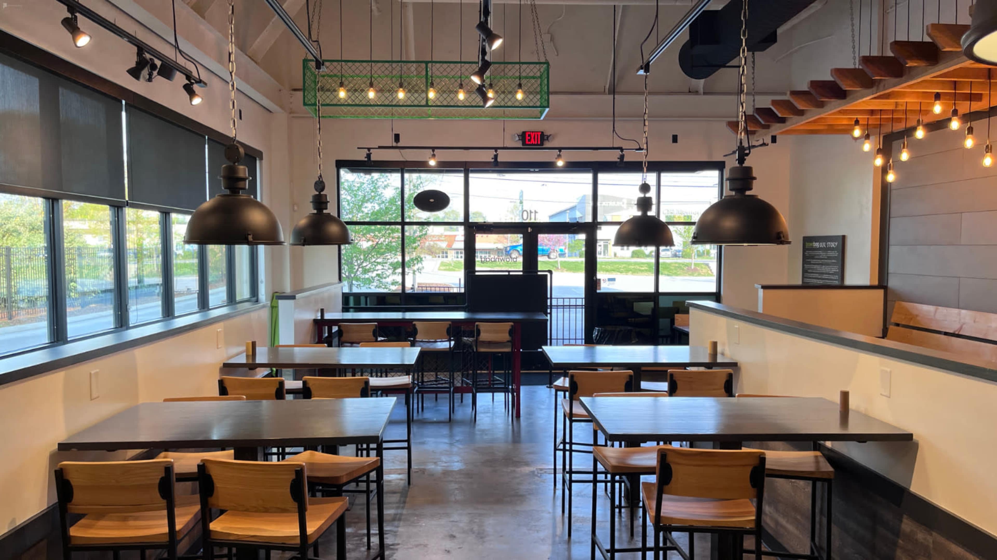 A modern restaurant interior with a series of wooden tables and chairs, large pendant lights, and a glass entrance door.