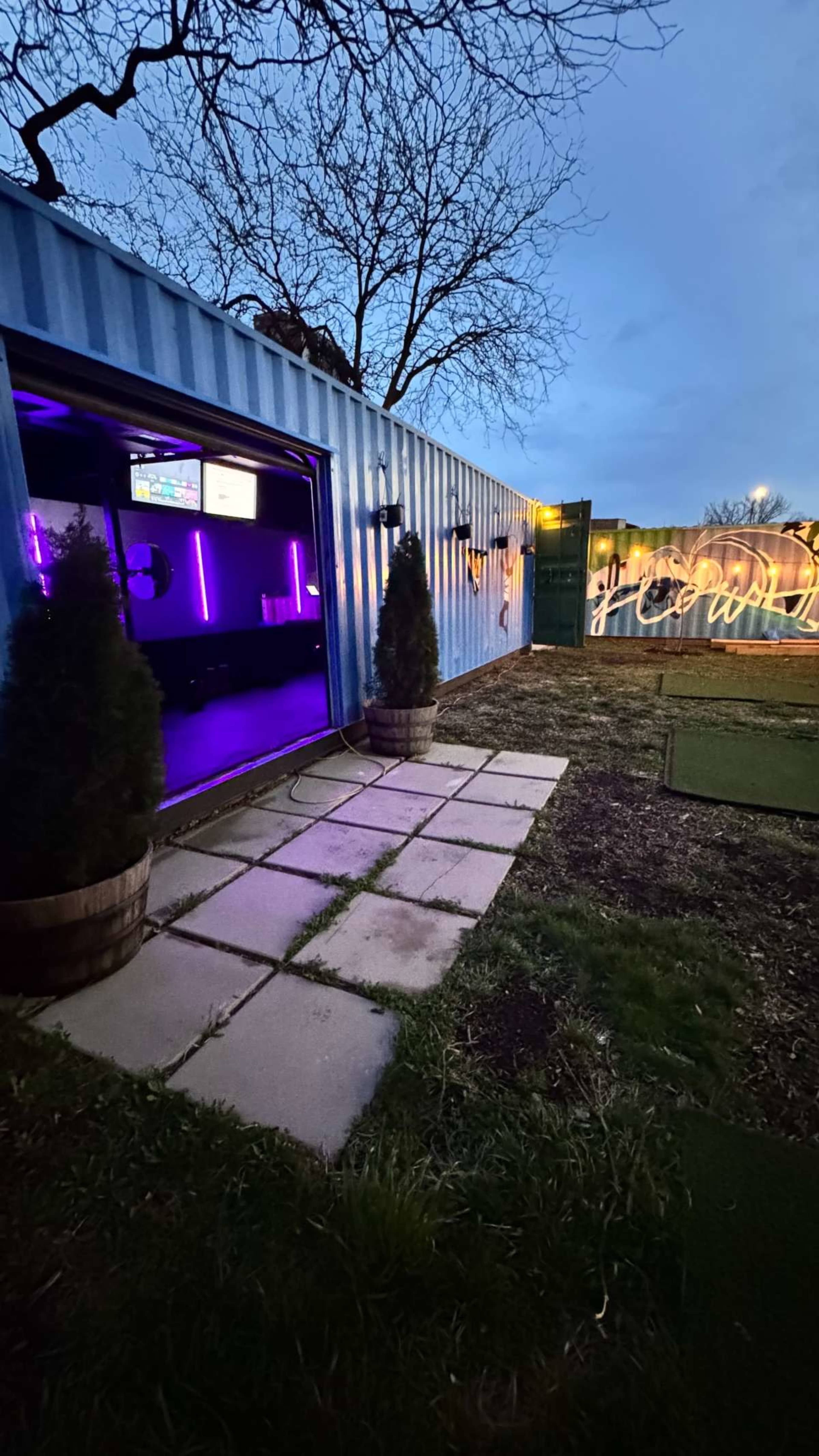 The image shows a shipping container at dusk, featuring a doorway illuminated by purple lights and two small trees in planters beside a stone pathway.