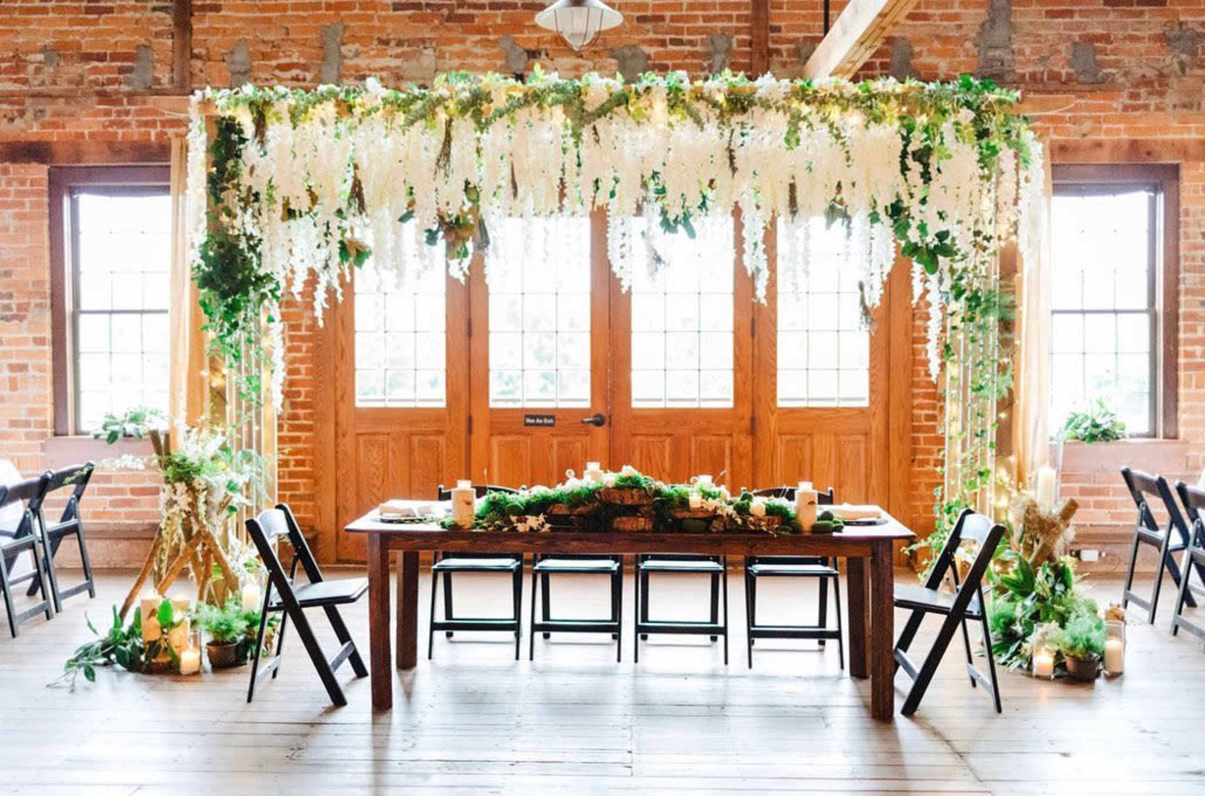 A decorated dining area features a long wooden table surrounded by black chairs, with a backdrop of greenery and floral arrangements against brick walls and double wooden doors.