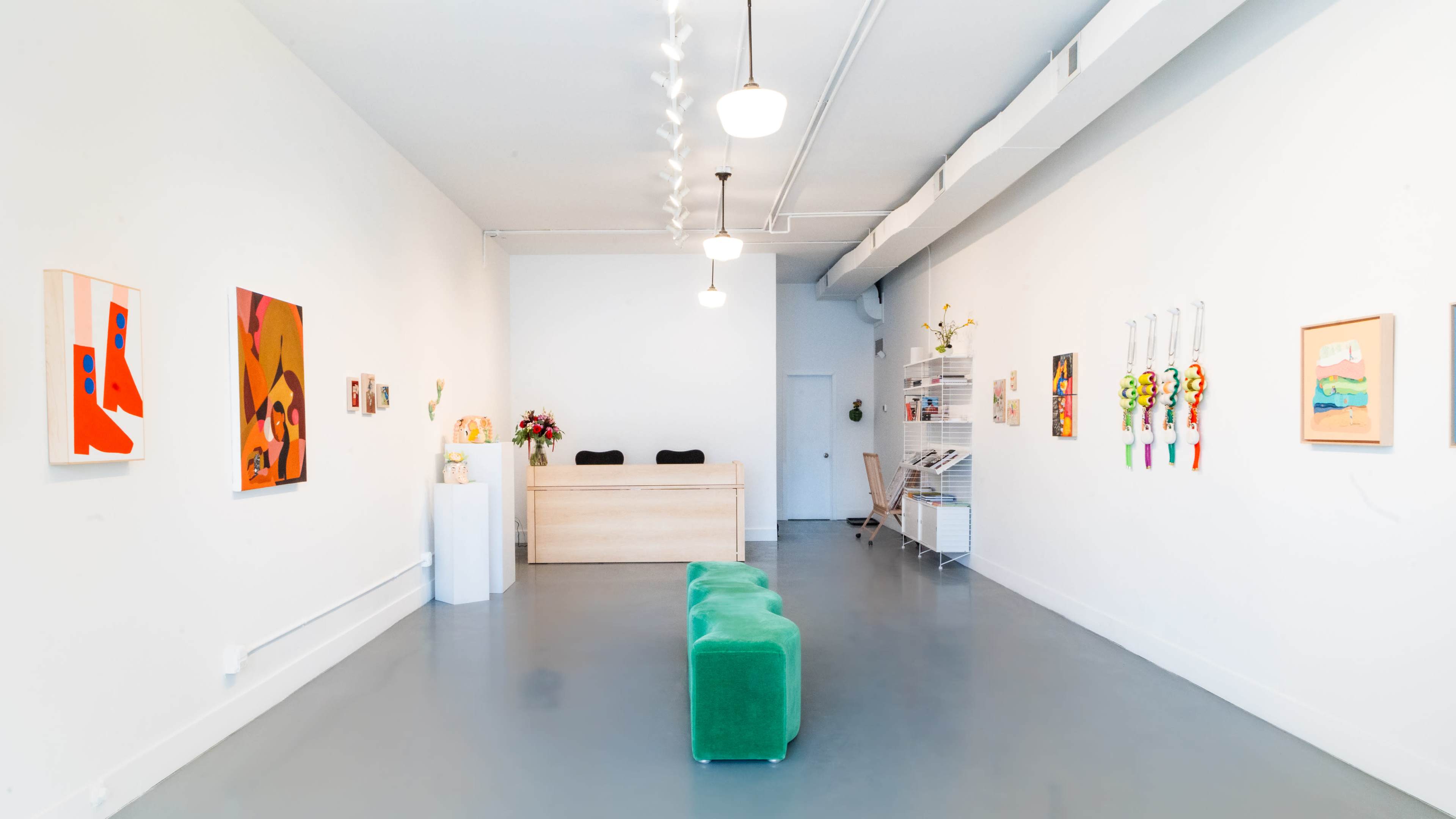 The image shows a bright, minimalist art gallery with a reception desk, colorful paintings on the walls, and a green seating area in the center.