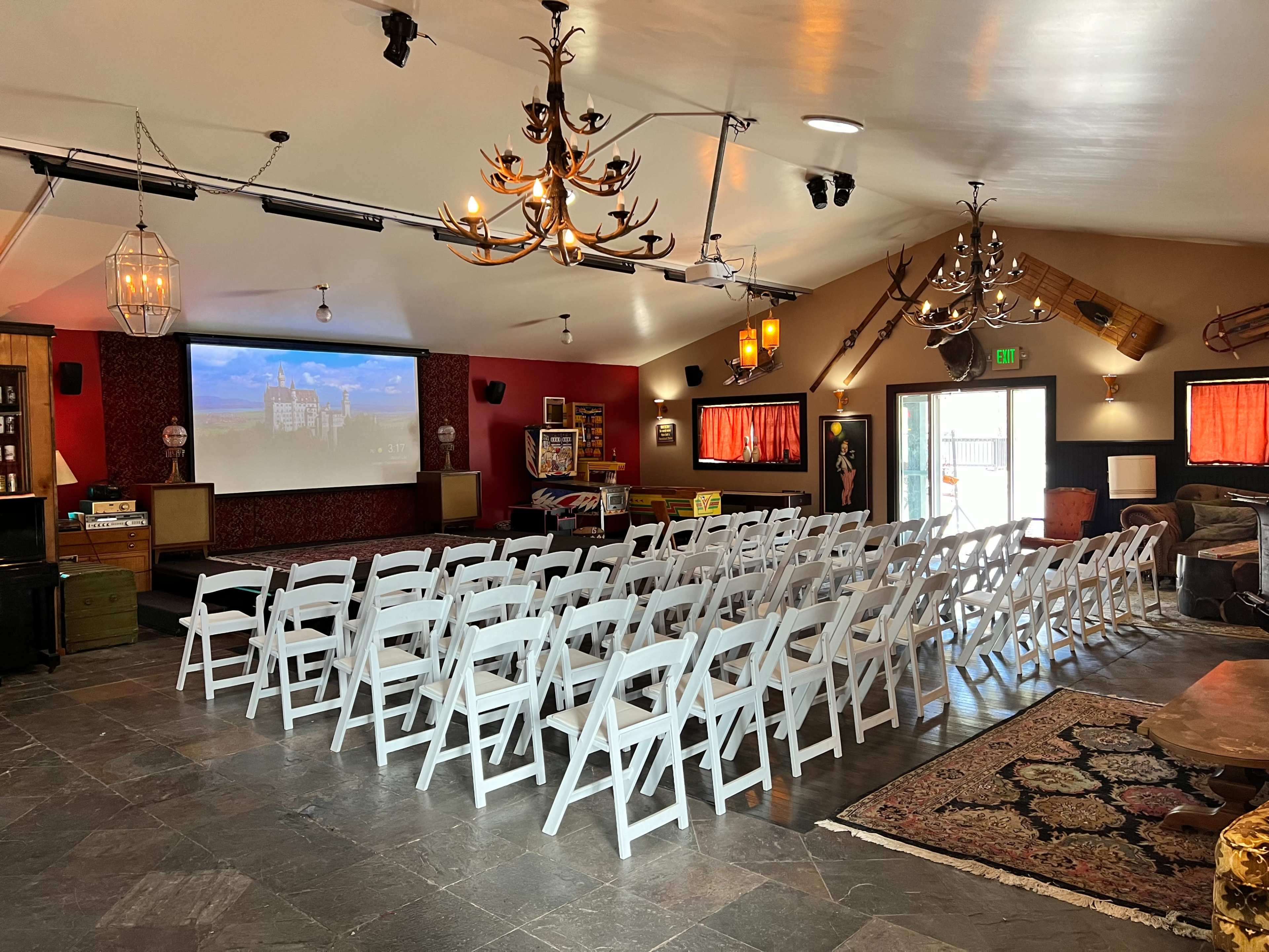 A spacious indoor venue set up with rows of white folding chairs facing a large projection screen, with chandeliers and decorative elements on the walls.