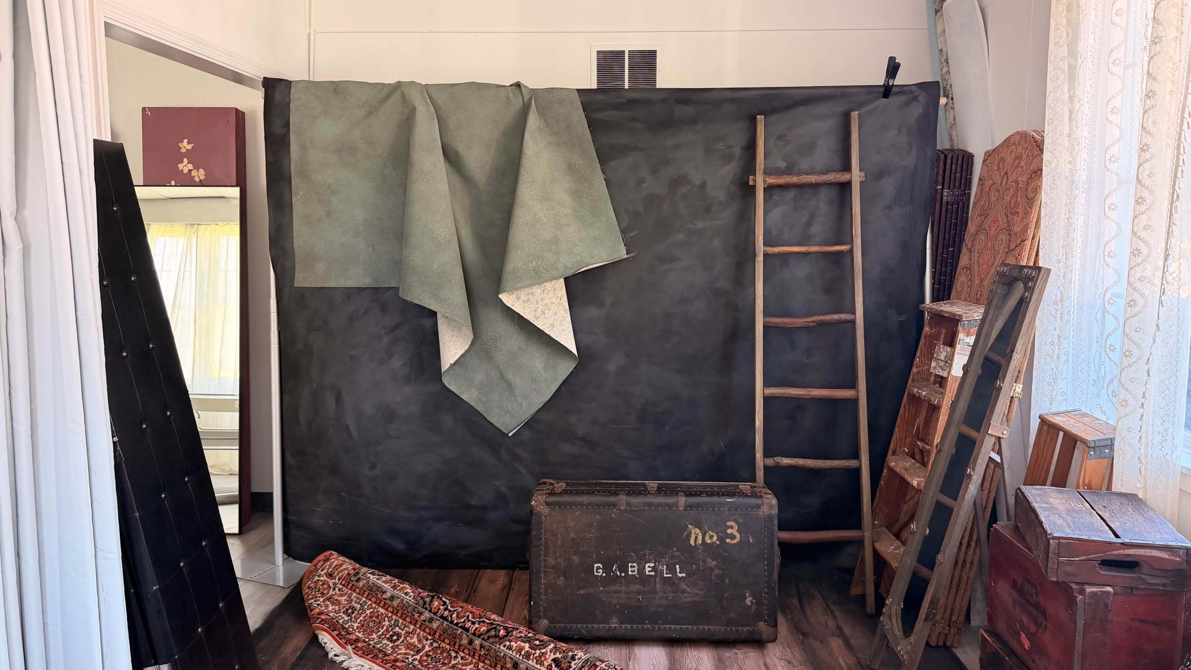 The image shows an indoor setting with a black backdrop, a green fabric draped over it, a wooden ladder against the wall, and various pieces of furniture, including an old trunk and rugs on the floor.