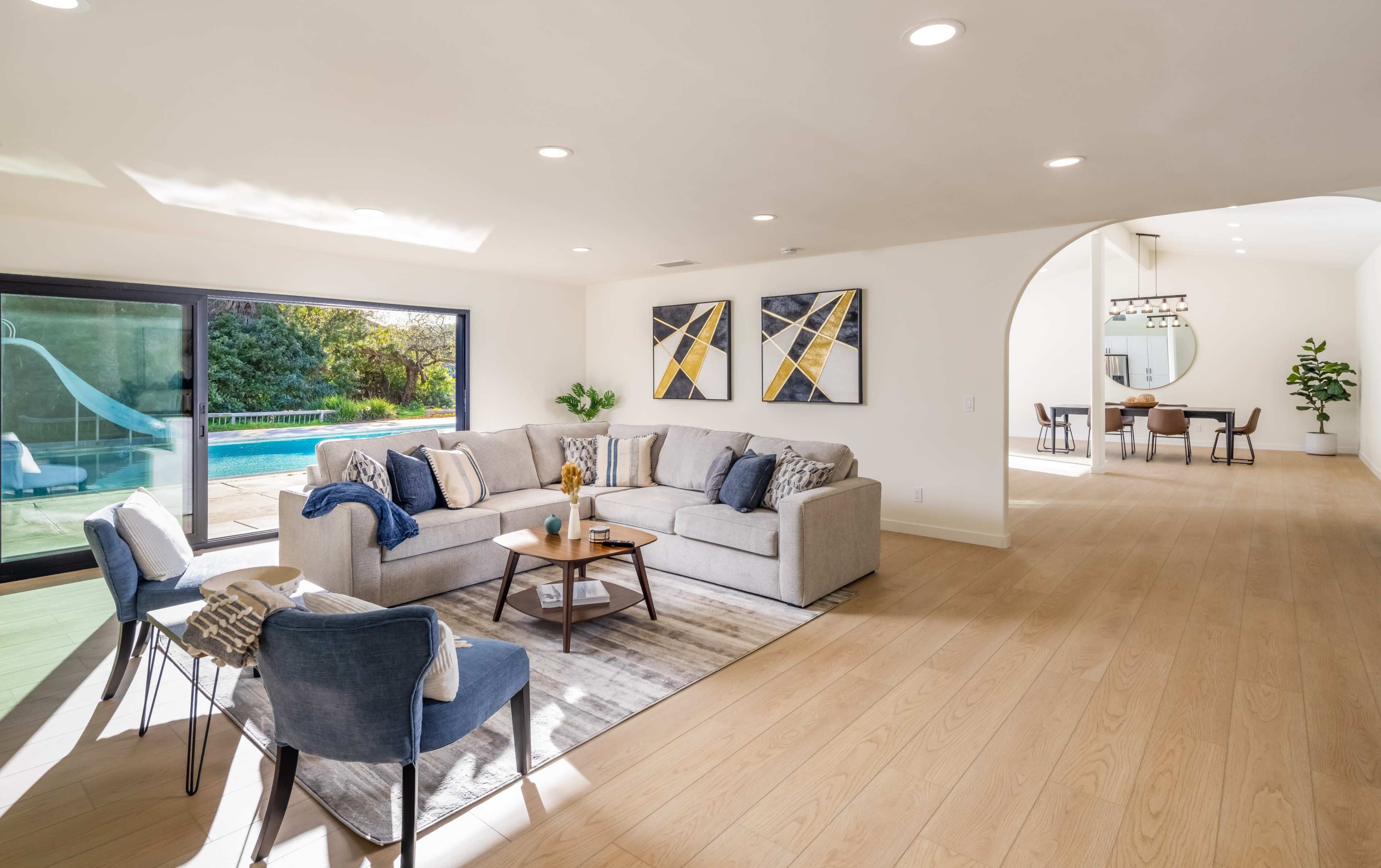 A spacious living room features a light-colored sectional sofa, two blue chairs, and large windows overlooking a pool area.