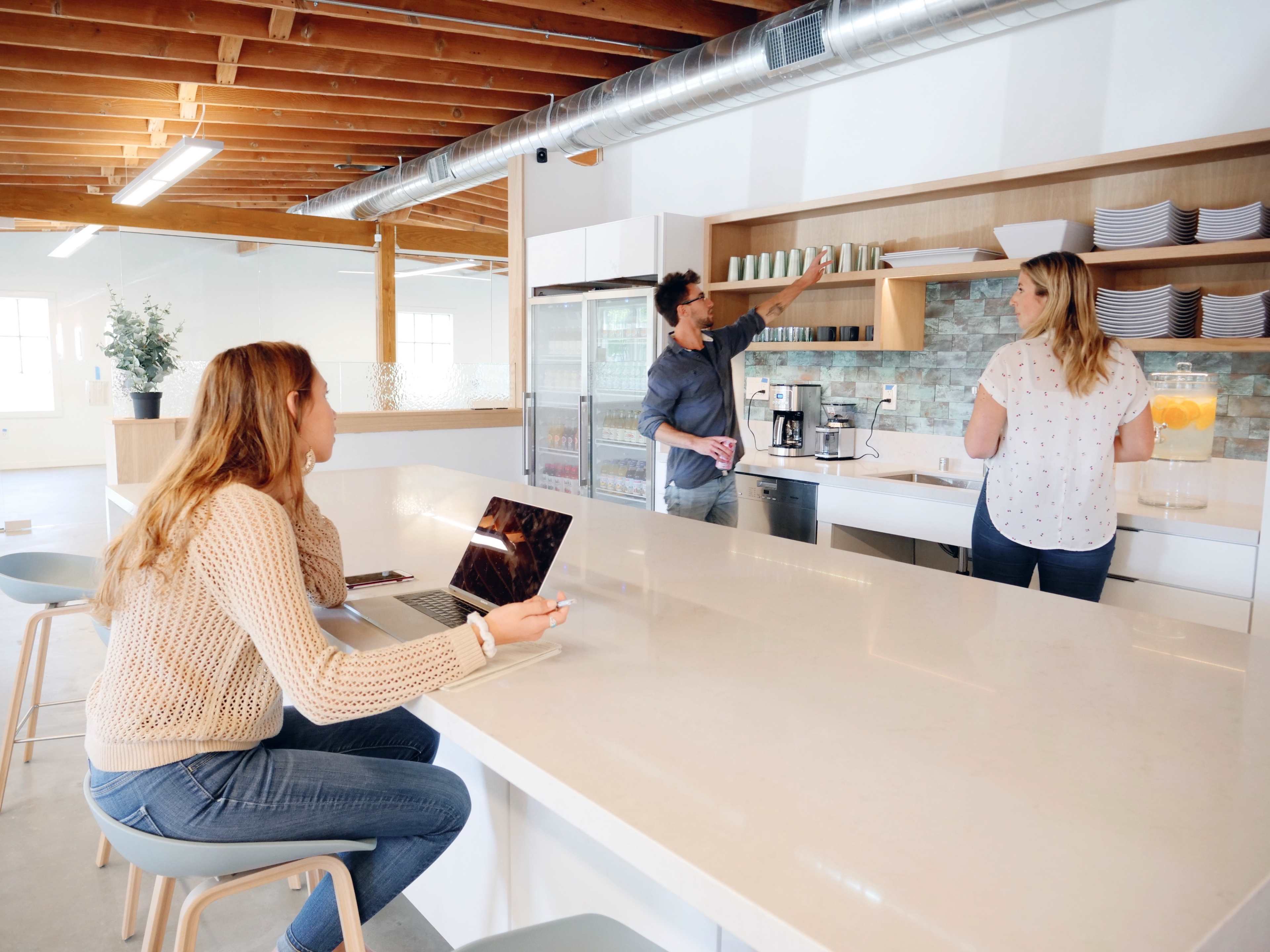 A man arranges cups on a shelf while a woman stands nearby, and another woman sits at a table with a laptop in a modern kitchen space.