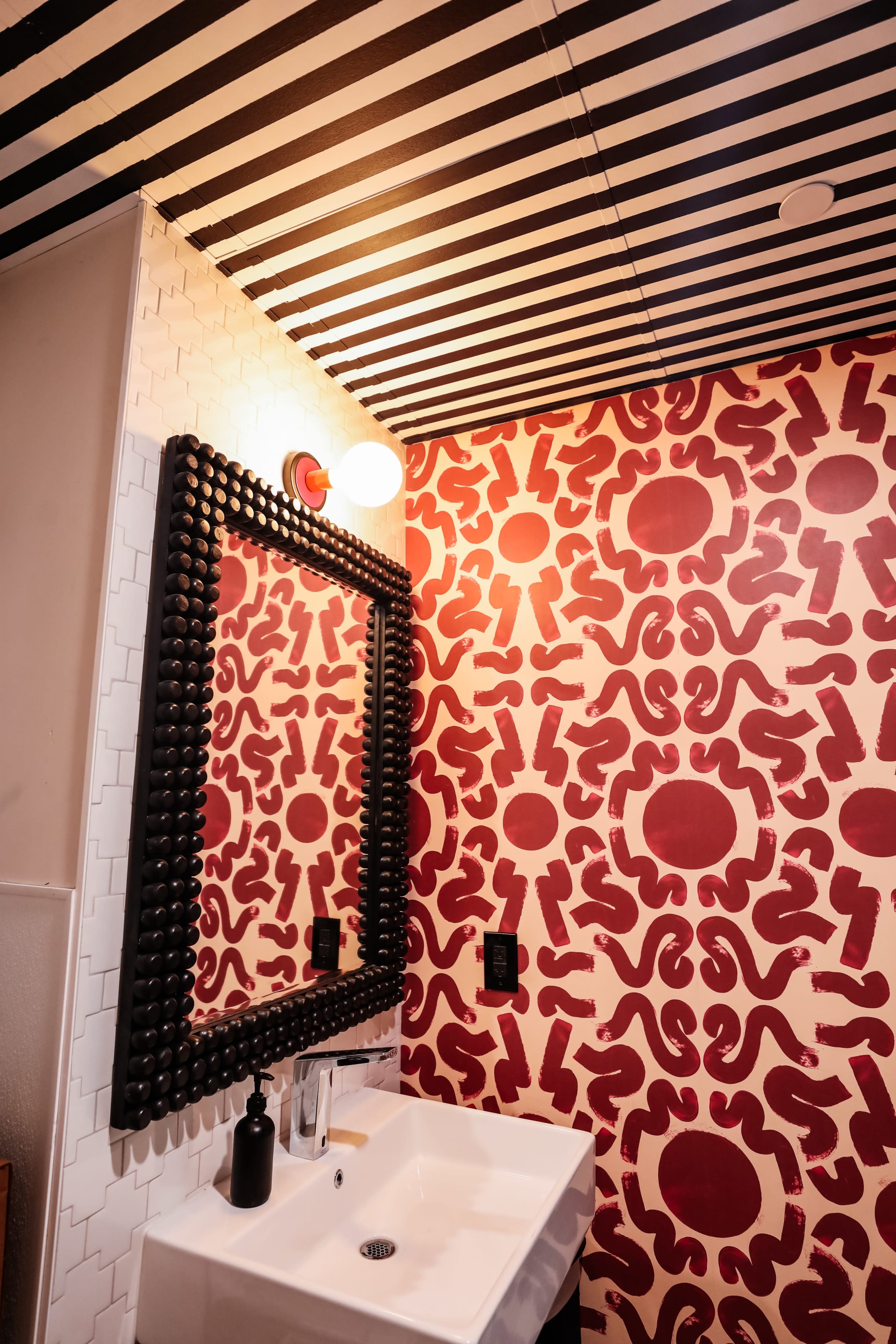 The image shows a bathroom with a patterned red and white wall, a black mirror, a white sink, and a striped black and white ceiling.