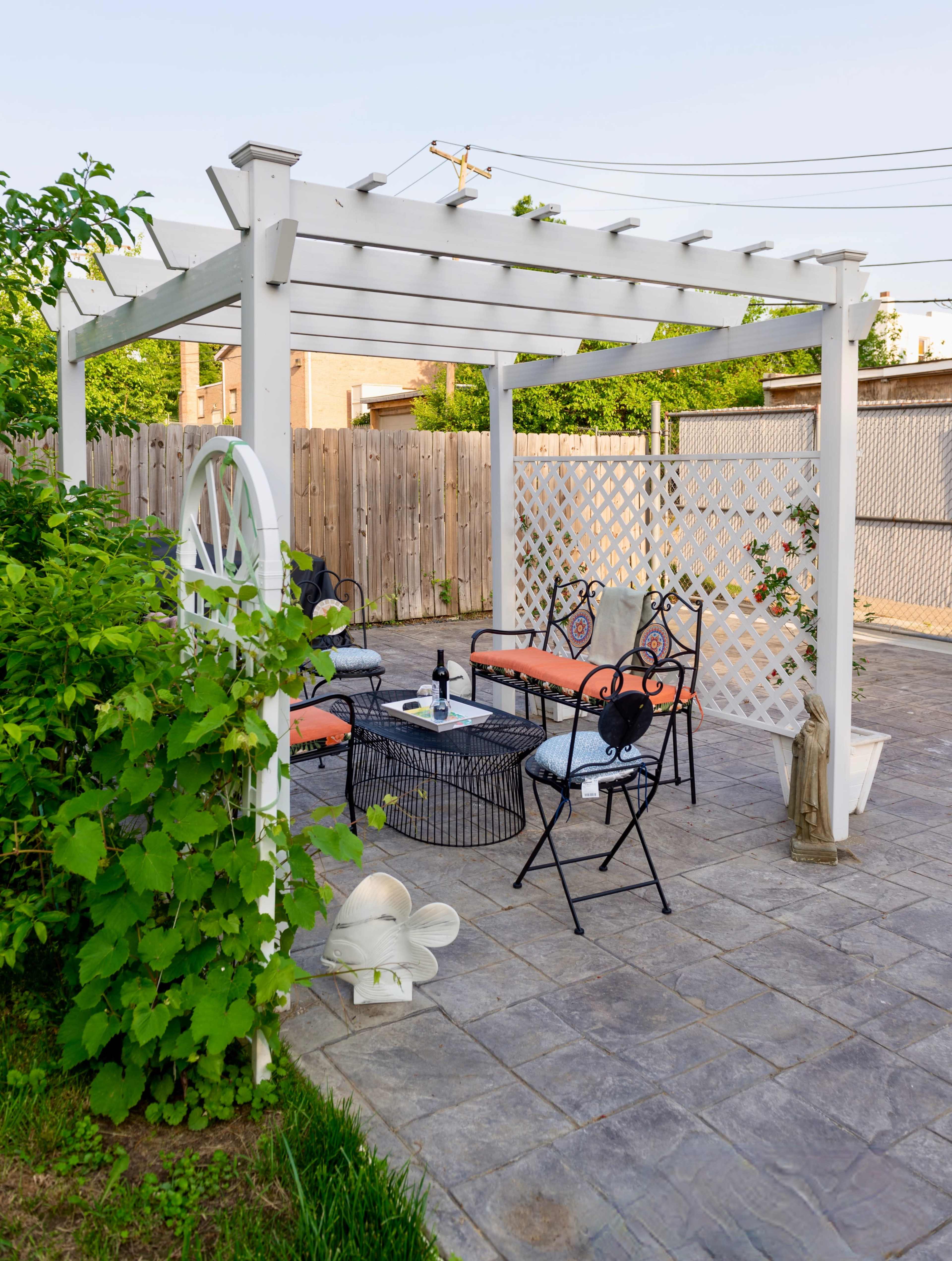 A shaded outdoor seating area features a white pergola with black metal chairs and a circular table, surrounded by greenery and a privacy screen.