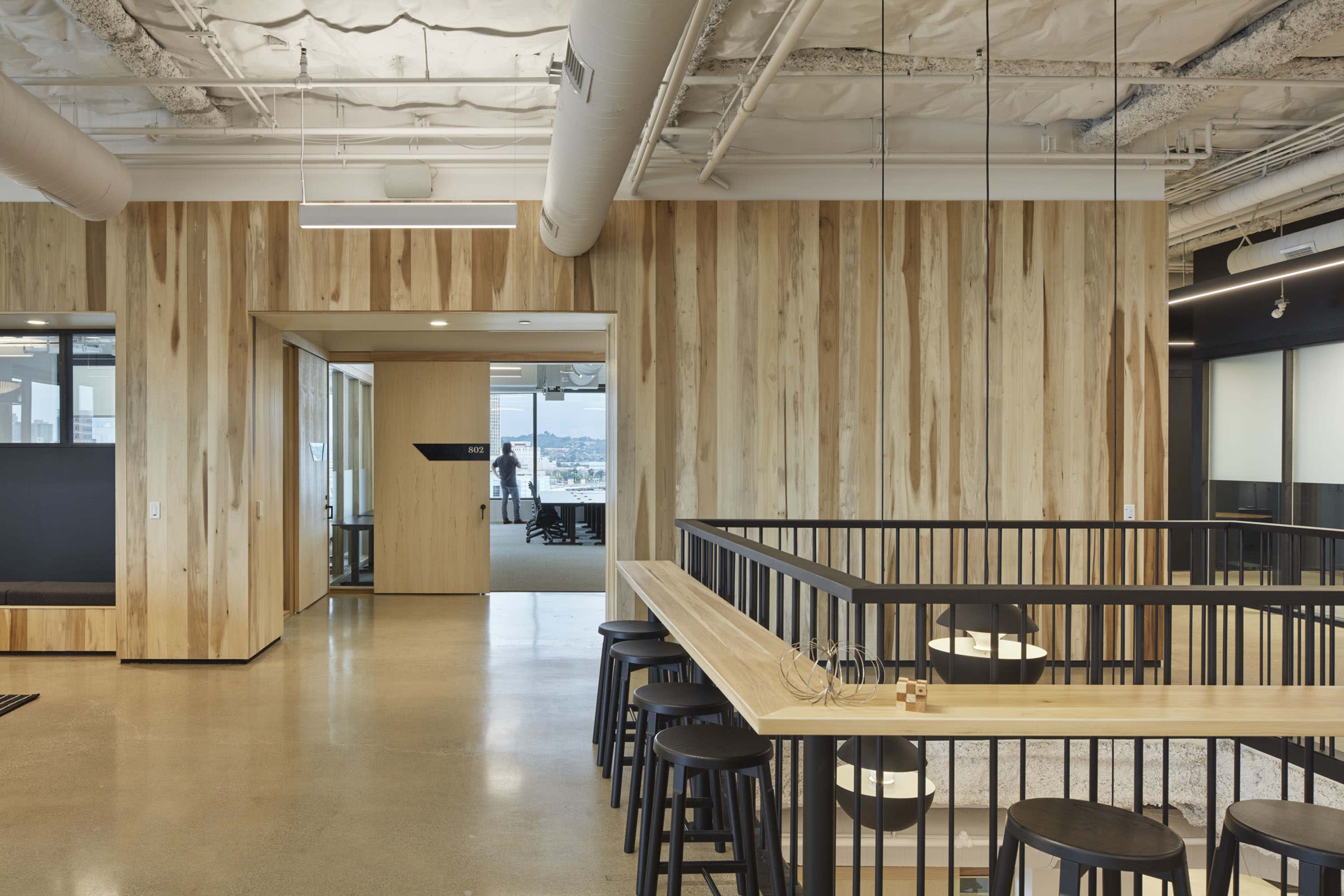 A modern office space featuring wooden walls, a low balcony area with black stools, and large windows allowing natural light to enter.