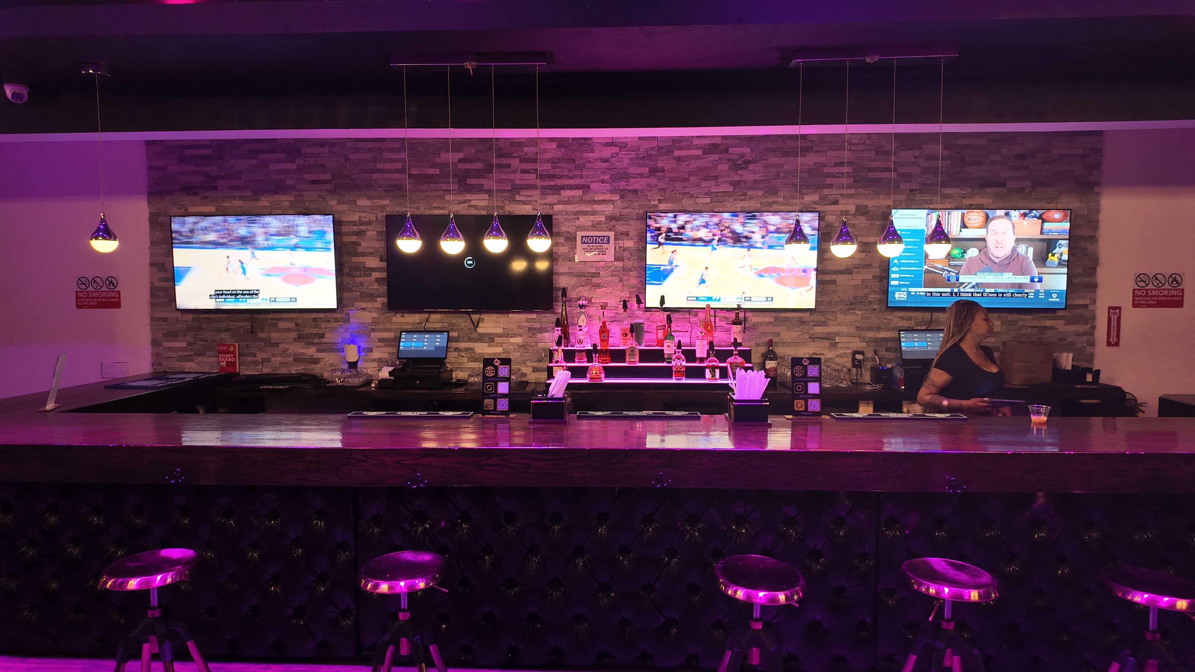 The image shows a modern bar area featuring multiple televisions on the wall, a long wooden counter, and bar stools, illuminated with purple lighting.