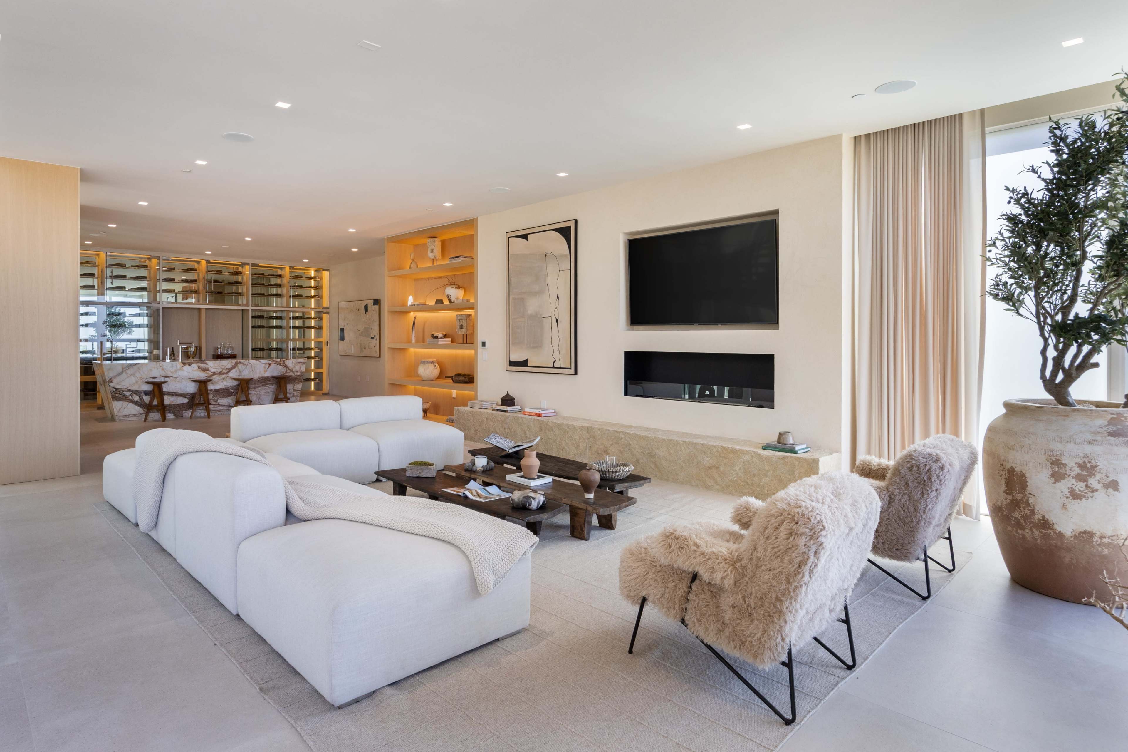 The image shows a modern living room featuring a white sectional sofa, a wooden coffee table, two textured armchairs, and a wall-mounted TV, with large windows allowing natural light.