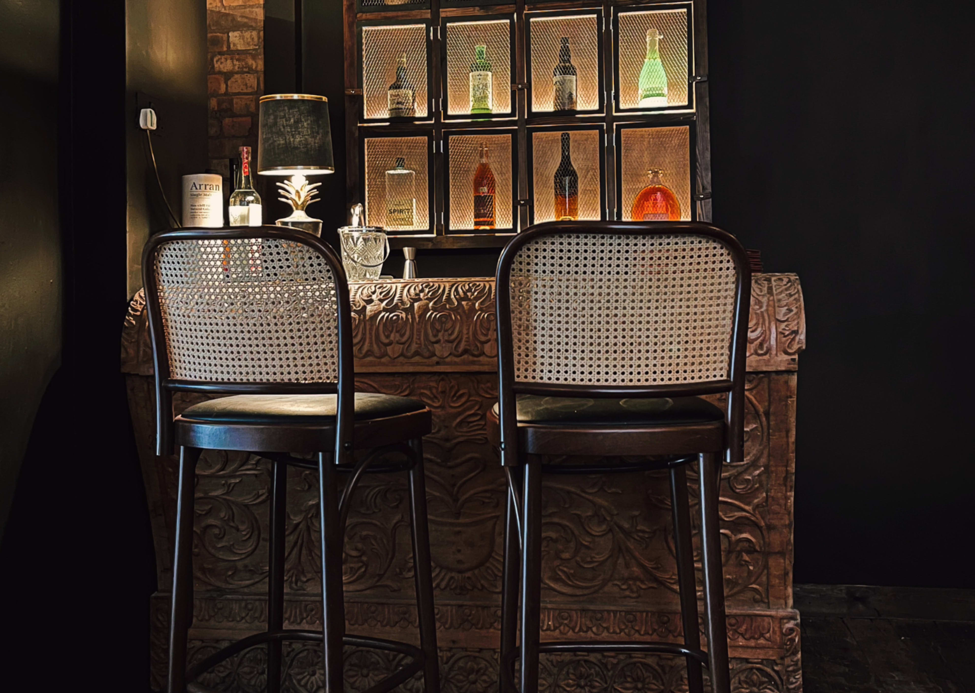 A dark bar area featuring two high-backed chairs in front of a decorative wooden counter adorned with bottles illuminated from behind.