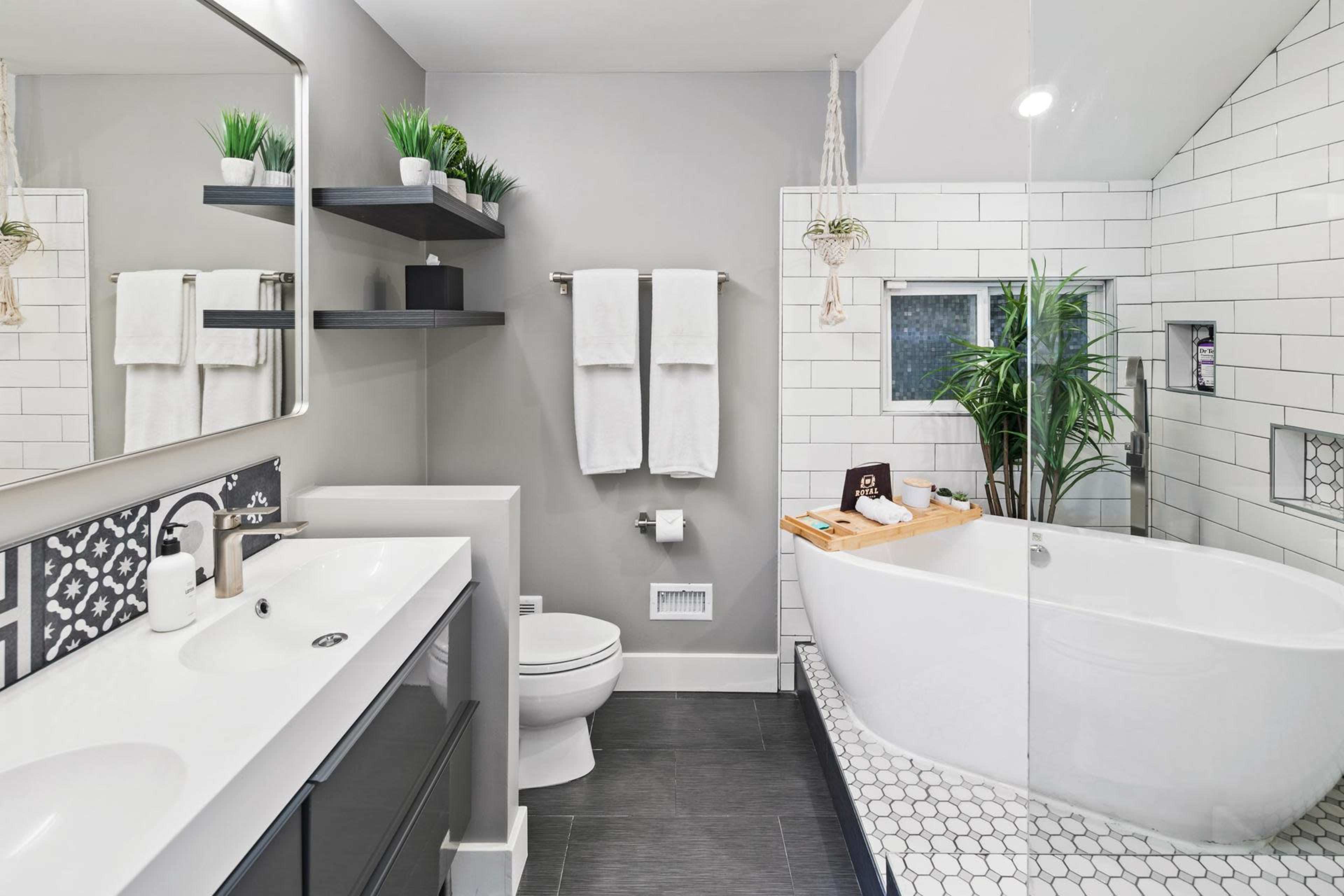 The bathroom features a white tiled shower with a glass enclosure, a freestanding bathtub, a double sink vanity, and wall-mounted shelves with plants.