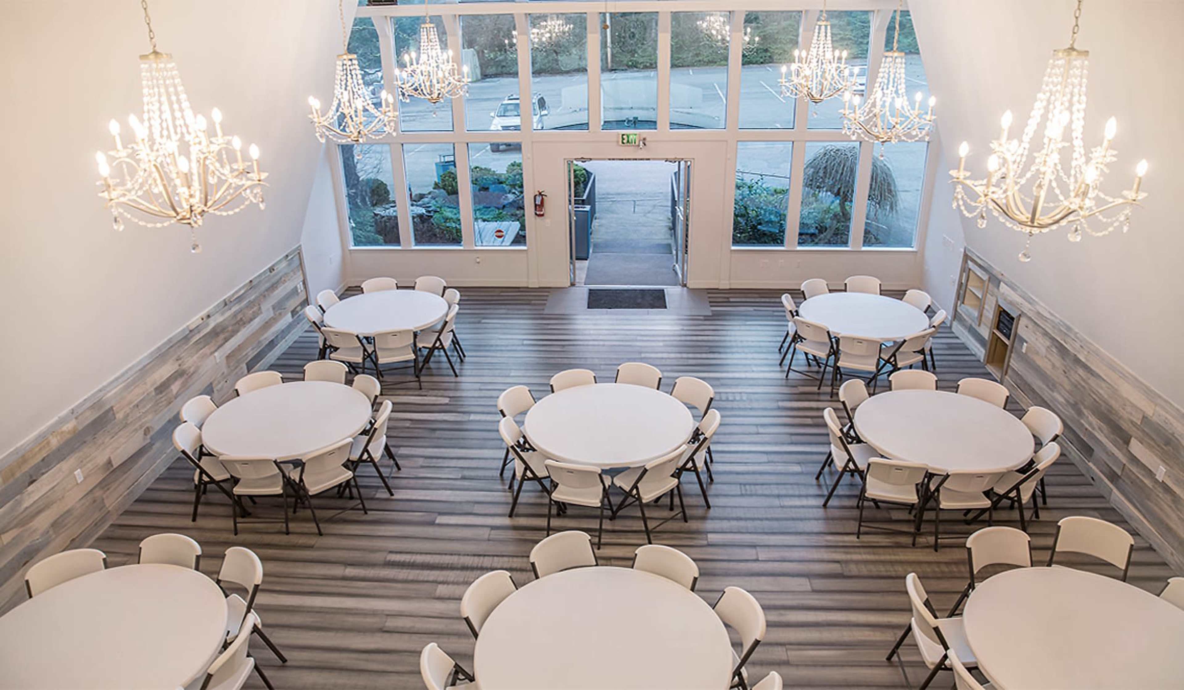 A spacious banquet room with round tables set up and large chandeliers hanging from the ceiling, illuminated by natural light from large windows.