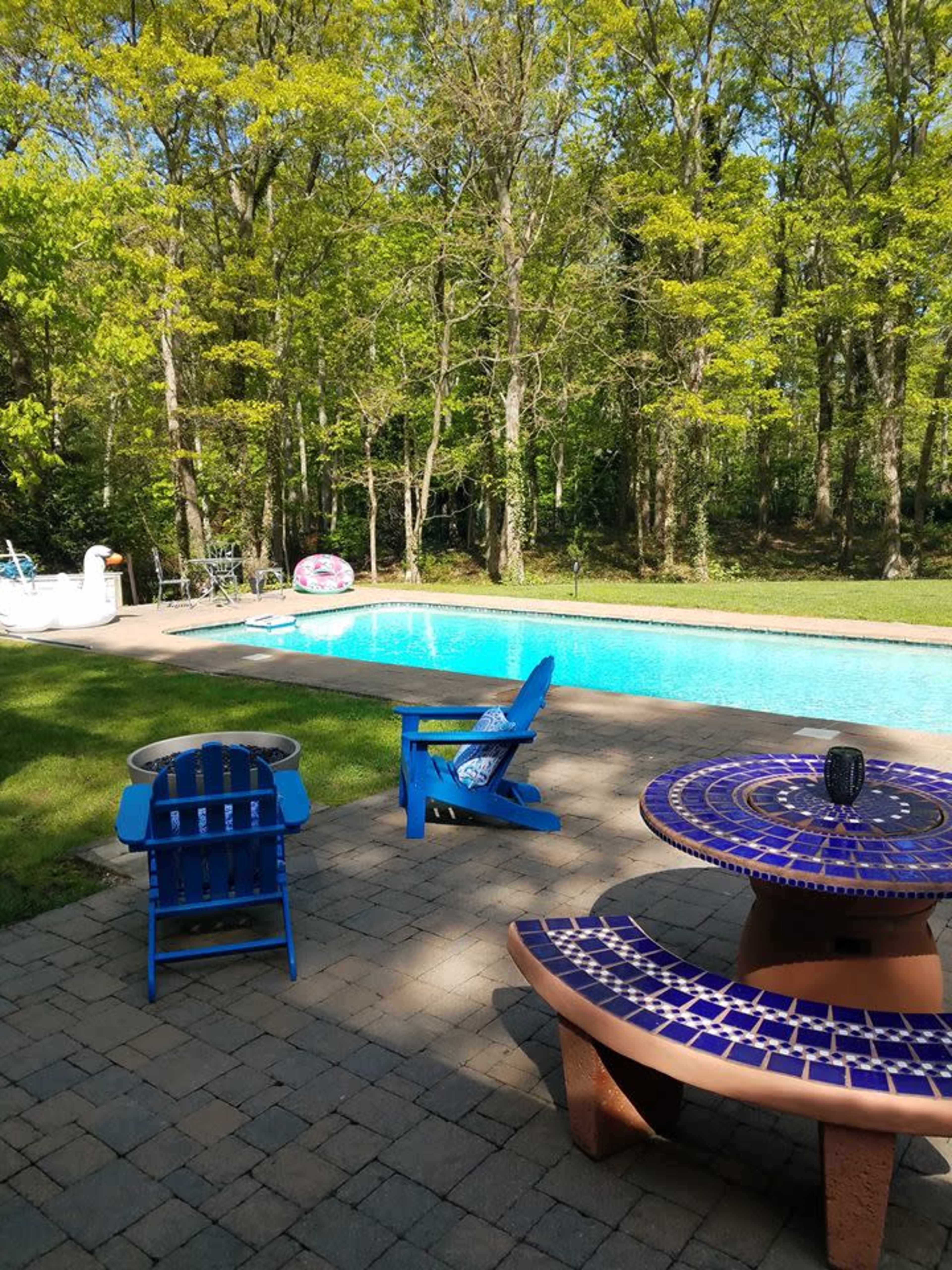 A backyard scene features a blue pool surrounded by trees, with colorful patio furniture and a stone patio.