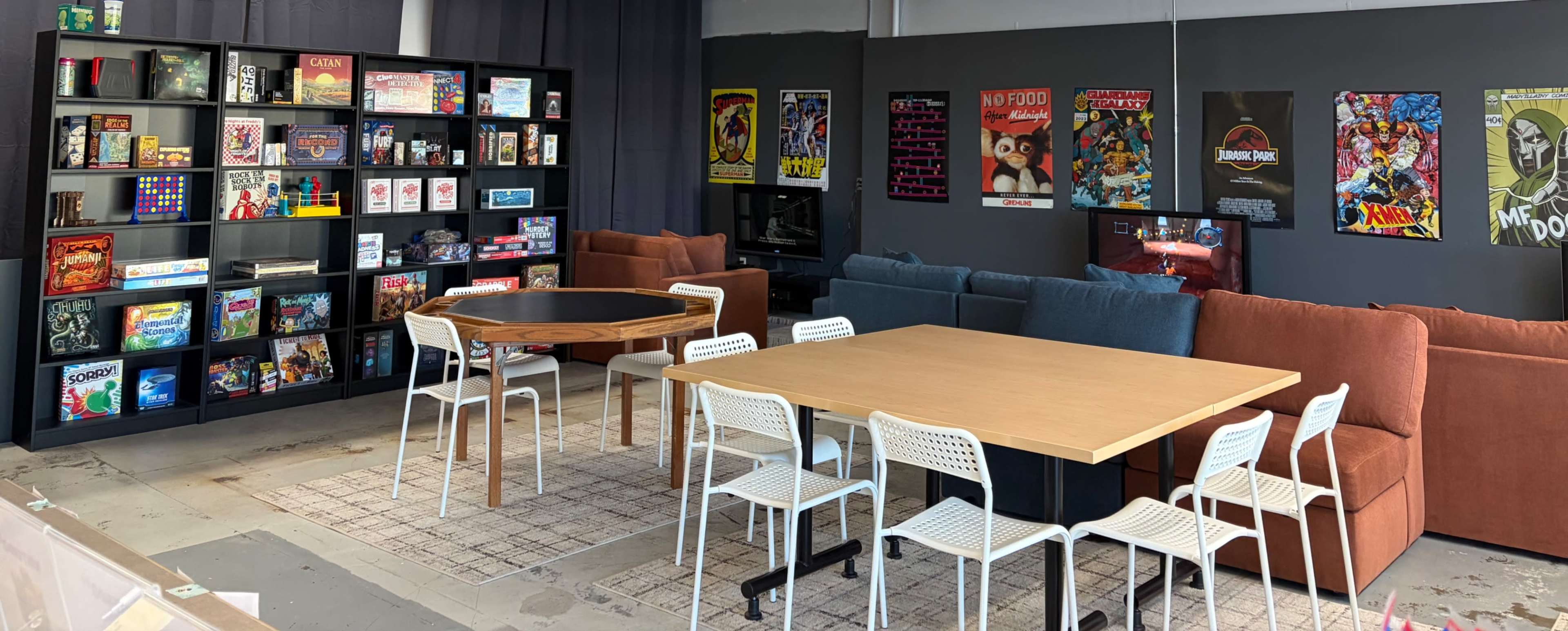 The image shows a space featuring shelves filled with board games, a circular wooden table surrounded by chairs, and a large rectangular table with white chairs, all set against a backdrop of colorful posters and a couch seating area.