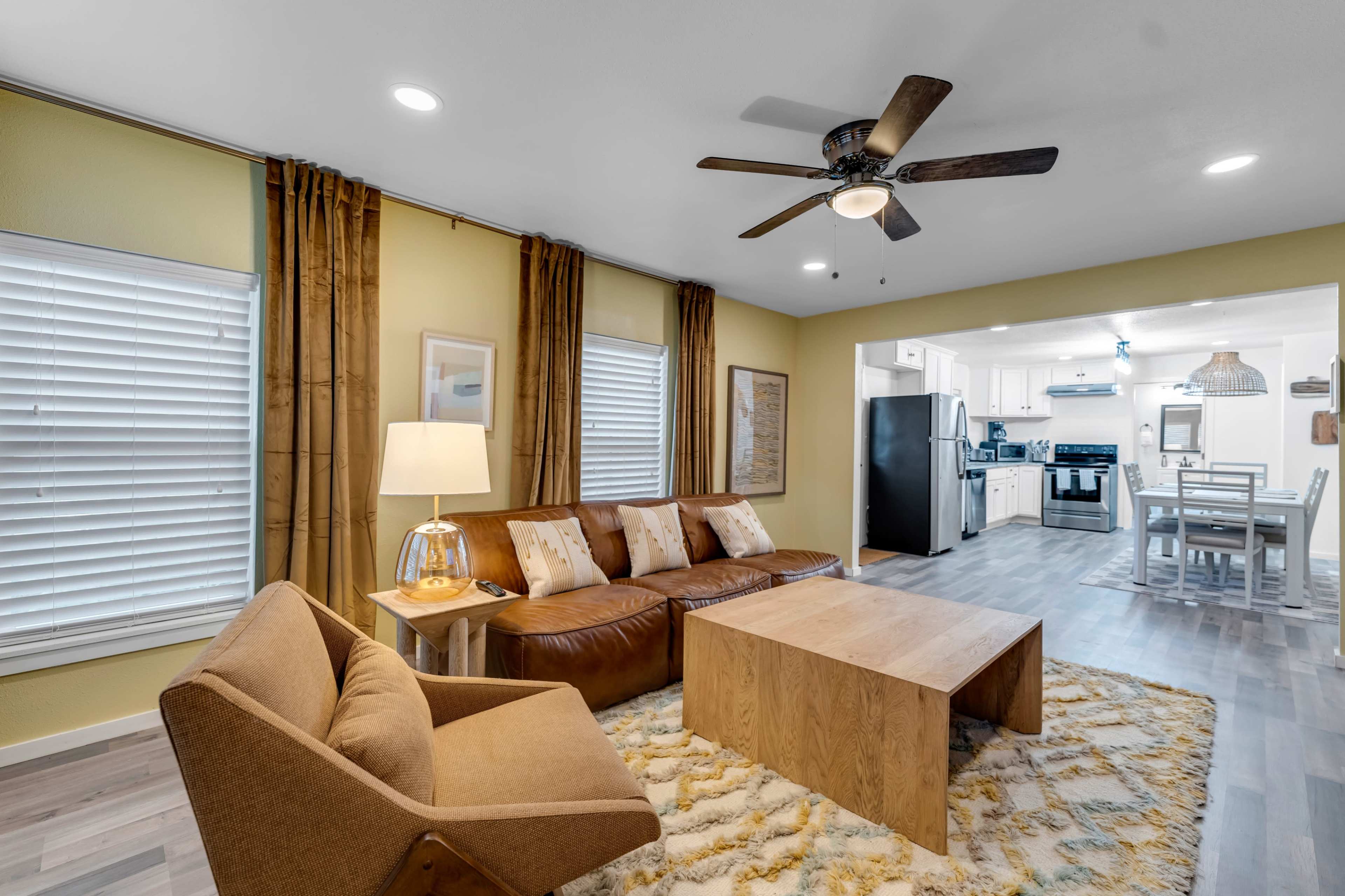 The image shows a modern living room with a brown leather sofa, a wooden coffee table, and an armchair, leading into a kitchen area illuminated by pendant lights.