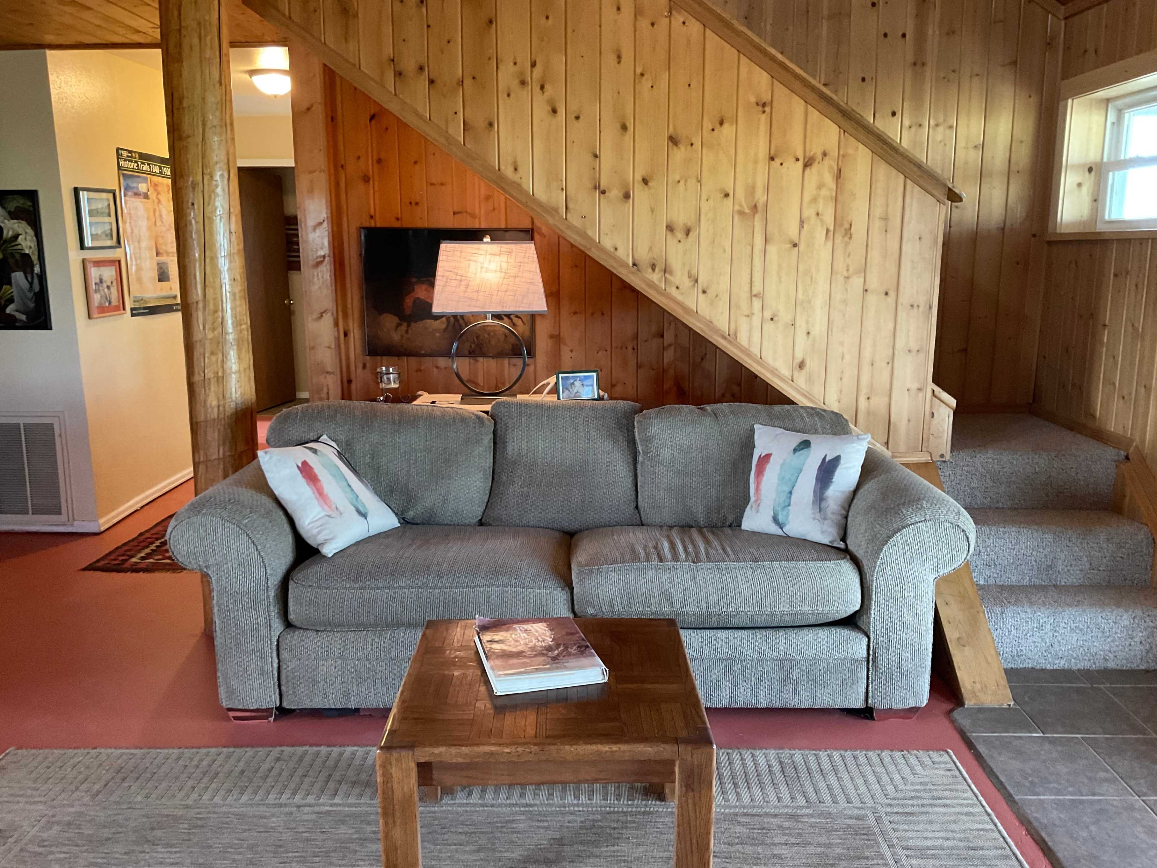 The image shows a living room with a gray sofa, a wooden coffee table, and a staircase leading to the upper level, all surrounded by wooden paneling.