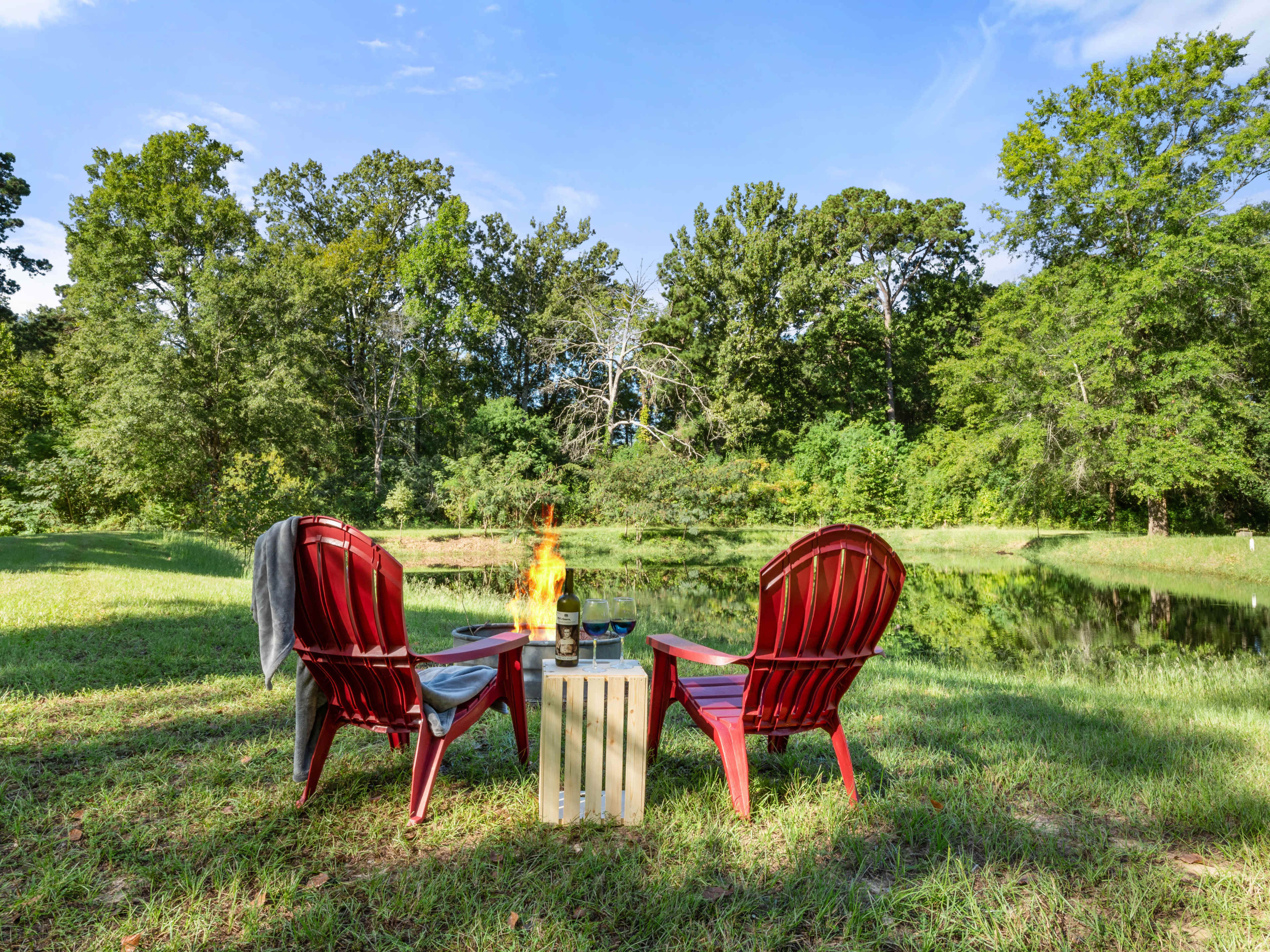 Two red Adirondack chairs face a small fire pit beside a pond, with a table holding glasses in the foreground and trees lining the background.